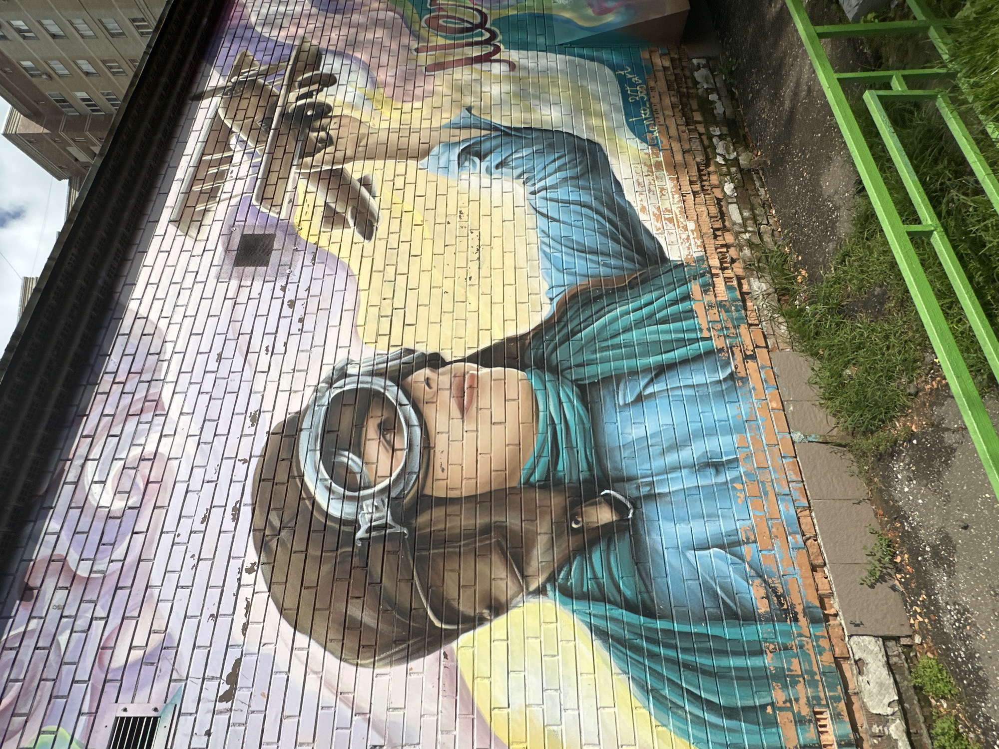 This is a photograph of a mural painted on a brick wall in the city of Podolsk, Russia. The mural depicts a person wearing a vintage aviator's helmet and goggles, with a blue scarf around their neck. The person's arm is extended, holding what appears to be a miniature airplane. The background of the mural transitions from purple and pink clouds at the top to a bright yellow sky. The brick wall itself shows signs of wear and tear, with some bricks chipped and mortar missing. In the foreground, there is green grass and a section of a green metal fence. In the upper right portion of the image, parts of residential buildings are visible. The mural is signed with what appears to be "Sanko 365 art" and "2022".