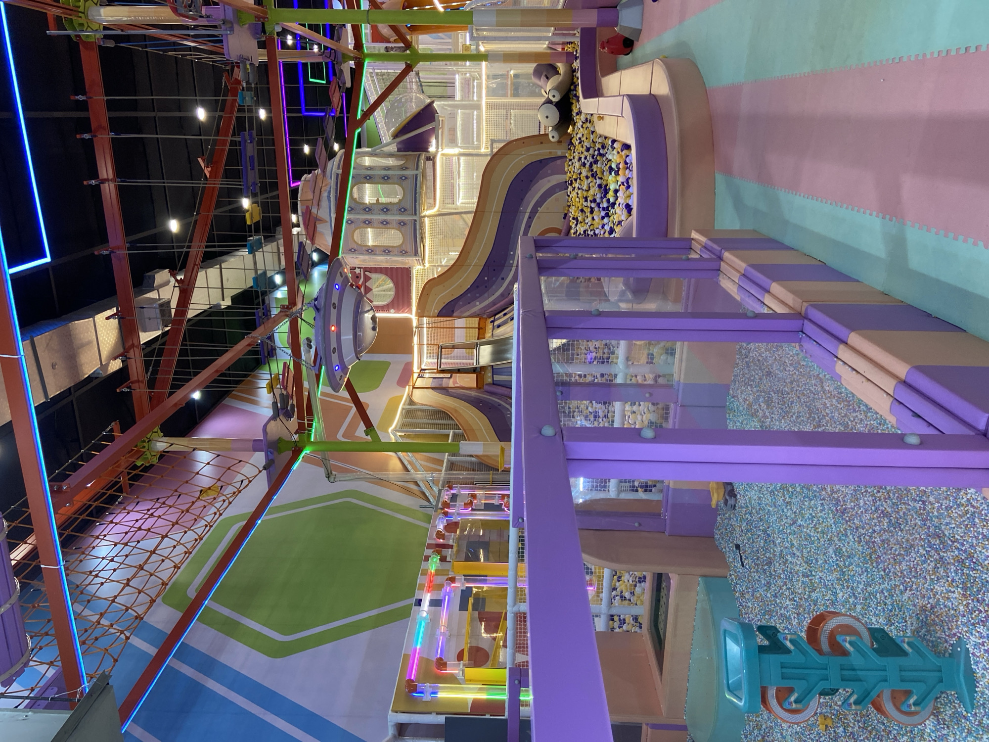 The image depicts an indoor children's play area, likely a soft play center or arcade. The environment is brightly colored with a predominant palette of purple, pink, green, and blue. There are several features characteristic of such a venue: a ball pit filled with multi-colored balls, a large, multi-colored slide with a wavy design, a rope net climbing structure suspended from the ceiling, and various tunnels and tubes. In the foreground, a low purple barrier encloses a section of the play area, and to the left, a teal-colored vertical play structure with rotating orange wheels is visible. In the background, a large, UFO-shaped play structure is suspended. The overall impression is one of a lively and engaging space designed for children's entertainment. This location is identified as being in Kecamatan Pati, Indonesia.