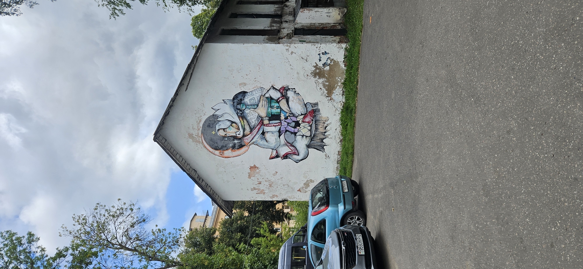 The image shows a mural painted on the side of a weathered, white building. The mural depicts a figure with a headdress, holding a turquoise object, against a backdrop of a crescent moon and stars. The building itself appears aged, with peeling paint and dark, recessed doorways on the right side. In front of the building, a gray car and a blue hatchback are parked on an asphalt surface. Green grass and trees are visible behind the cars and along the base of the building. The sky is partly cloudy with patches of blue visible. The location is the city of Vitsebsk, Belarus.