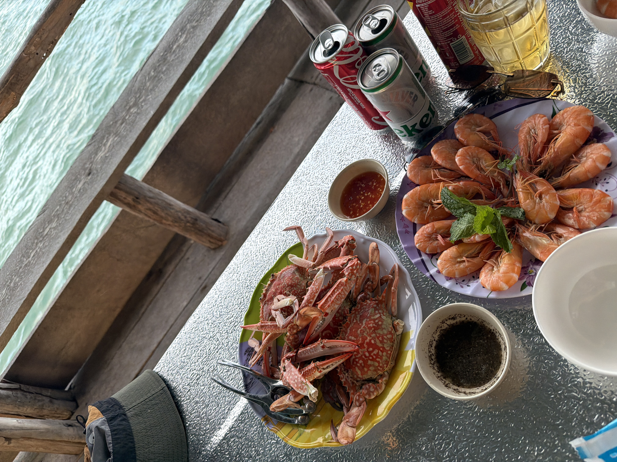 This image, likely taken in Ninh Ích, Vietnam, depicts a table laden with seafood. Specifically, there are crabs and shrimp arranged on separate plates. Condiments are in small bowls. Beverages, including Coca-Cola and Heineken, along with a glass of beer, are also present. The scene is set on a textured metal table, likely outdoors, with a view of the sea visible through a wooden structure. A green cap is visible in the lower left corner. There are no people visible in the image.