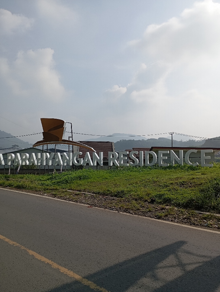 The image displays a static outdoor scene in Pataruman, Banjar, West Java, Indonesia, under daylight conditions with a partly cloudy sky. The foreground features a dry asphalt road, occupying approximately the lower third of the frame, exhibiting a single solid yellow line along its left edge and dark, elongated shadows cast from the left. A strip of unmanicured green grass, with visible dark soil patches, separates the road from a large, white, three-dimensional sign. This sign prominently spells "PARAHYANGAN RESIDENCE." To the left of the "PARAHYANGAN" text, a distinct brown, multi-curved architectural or sculptural element is visible. Directly behind the sign, a structure with a reddish-brown tiled roof is partially discernible. The background consists of multiple layers of undulating, verdant hills or mountains under the hazy sky. Overhead, power lines traverse the mid-ground, supported by at least one utility pole visible on the right side of the frame. No individuals are present, and no actions or interactions are depicted.