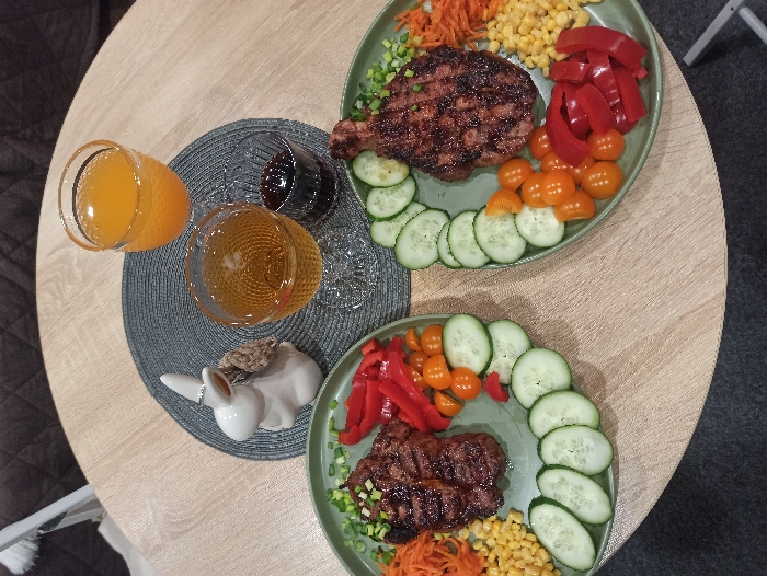 The scene depicts a static, indoor dining arrangement on a light wood-patterned round table in Nizhneye Zadolgoye, Russia. Two prepared meals are presented on sage-green oval plates. Each plate contains a central grilled meat item, garnished with chopped green onions, surrounded by sections of sliced cucumbers, orange cherry tomatoes, red bell pepper strips, shredded carrots, and yellow corn kernels.
Centered on a dark gray woven placemat between the plates are three beverage glasses. One glass, with a textured surface, holds an orange liquid. Another stemmed glass, also textured, contains a light amber liquid. A smaller dark glass with vertical ridges holds a dark liquid. To the left of the beverages stands a white ceramic rabbit figurine with a small, light brown woven accessory on its back and a silver ring on its left ear.
The background shows a dark gray or brown quilted upholstered surface, likely a sofa or bench, and a dark gray floor covering. No individuals are present, and no active actions or interactions are depicted.