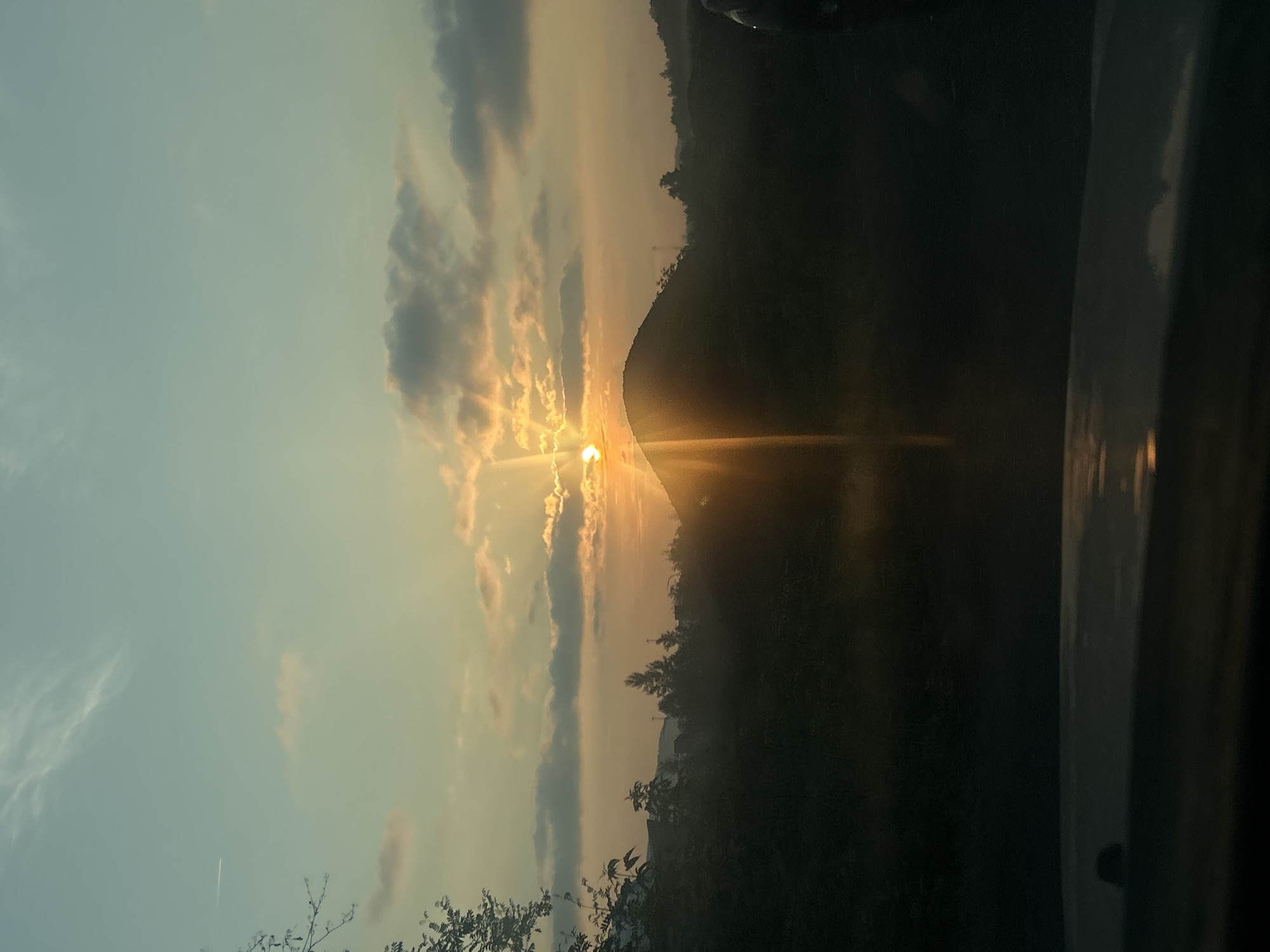 The image depicts a sunset over a silhouetted landscape. The sun, partially obscured by clouds and a rounded hill, casts bright rays of light across the sky. The sky is a gradient of light blue and pale yellow, with wispy clouds scattered throughout. Below the sky, a dark silhouette of trees and vegetation forms the foreground. The bottom of the image shows a dark, reflective surface, possibly a car's dashboard, with a subtle hint of the sunset's reflection. The overall scene is peaceful and serene, capturing the natural beauty of a sunset.