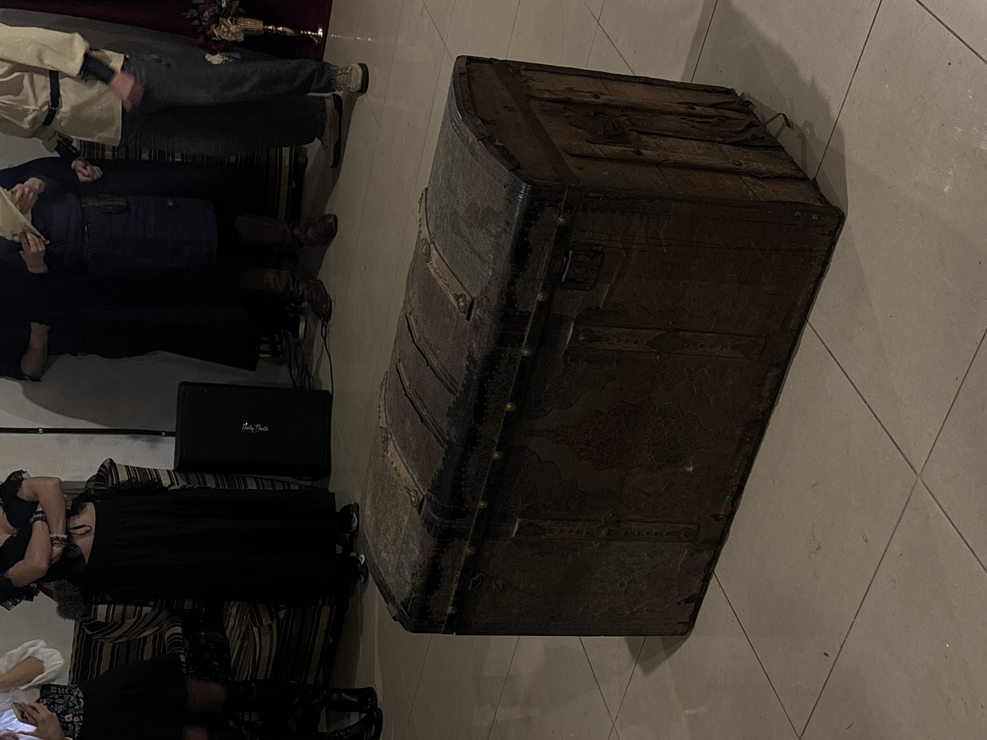 The image shows a cluttered indoor scene. In the foreground, a large, dark-colored, vintage steamer trunk dominates the frame. It is heavily detailed with metal accents and appears weathered. In the background, partially obscured by the trunk, are several individuals. To the left are two women sitting on a striped sofa. Near them, a speaker, with words Harley Benton on it, is also visible. Another person is seen in the upper left corner of the image. The environment has a tiled floor. The location cannot be identified as Varna, Bulgaria, based on the image.