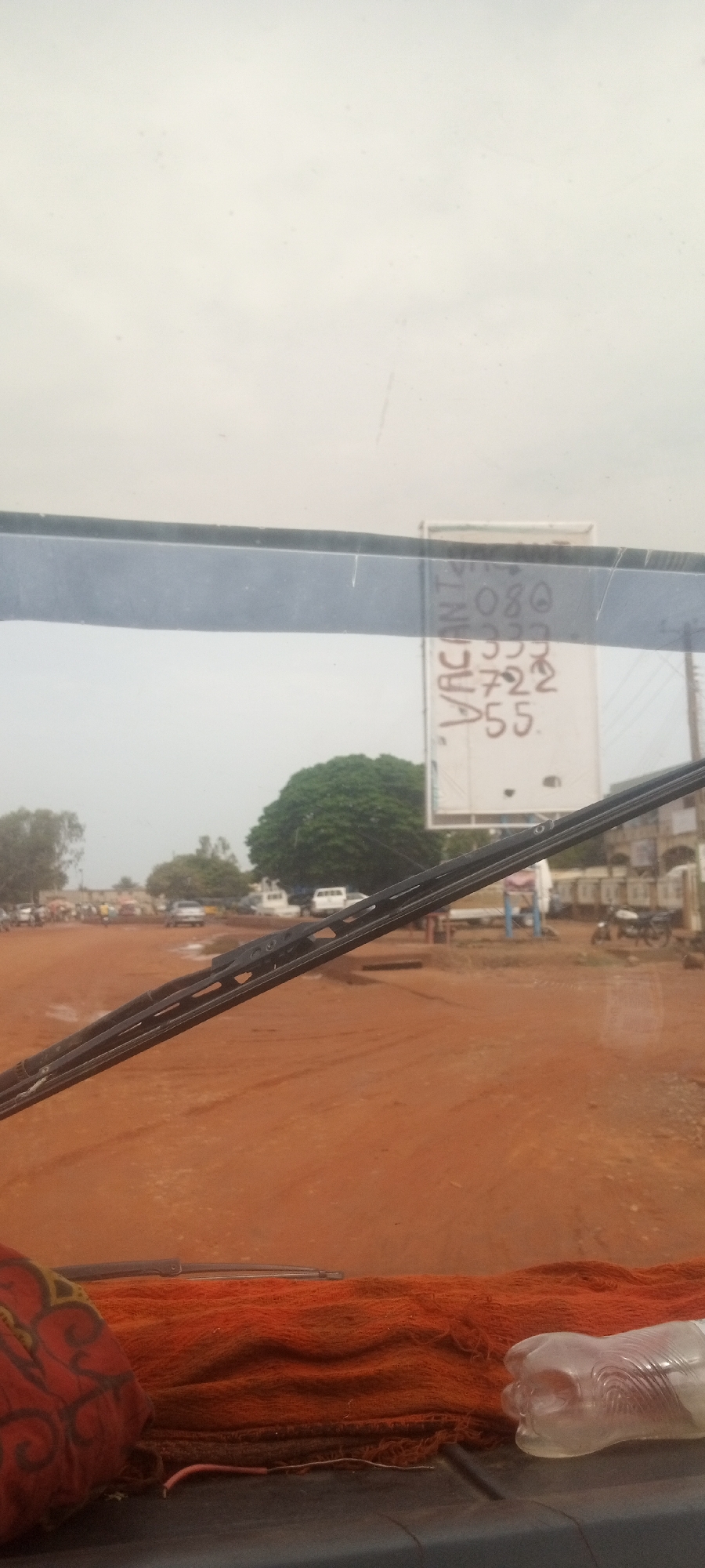 The image appears to be taken from inside a vehicle, looking out through the windshield. The scene shows a dusty, reddish-brown road in Barden Yana, Nigeria. In the background, there's a gathering of people, motorcycles, and vehicles, suggesting a populated area or possibly a marketplace. A large tree is visible in the middle distance. A sign with "VACANT" and some numbers (possibly a phone number) is partially visible on the right side of the frame. The windshield wipers are in the foreground, partially obscuring the view. A red patterned object and a clear plastic bottle are on the dashboard.