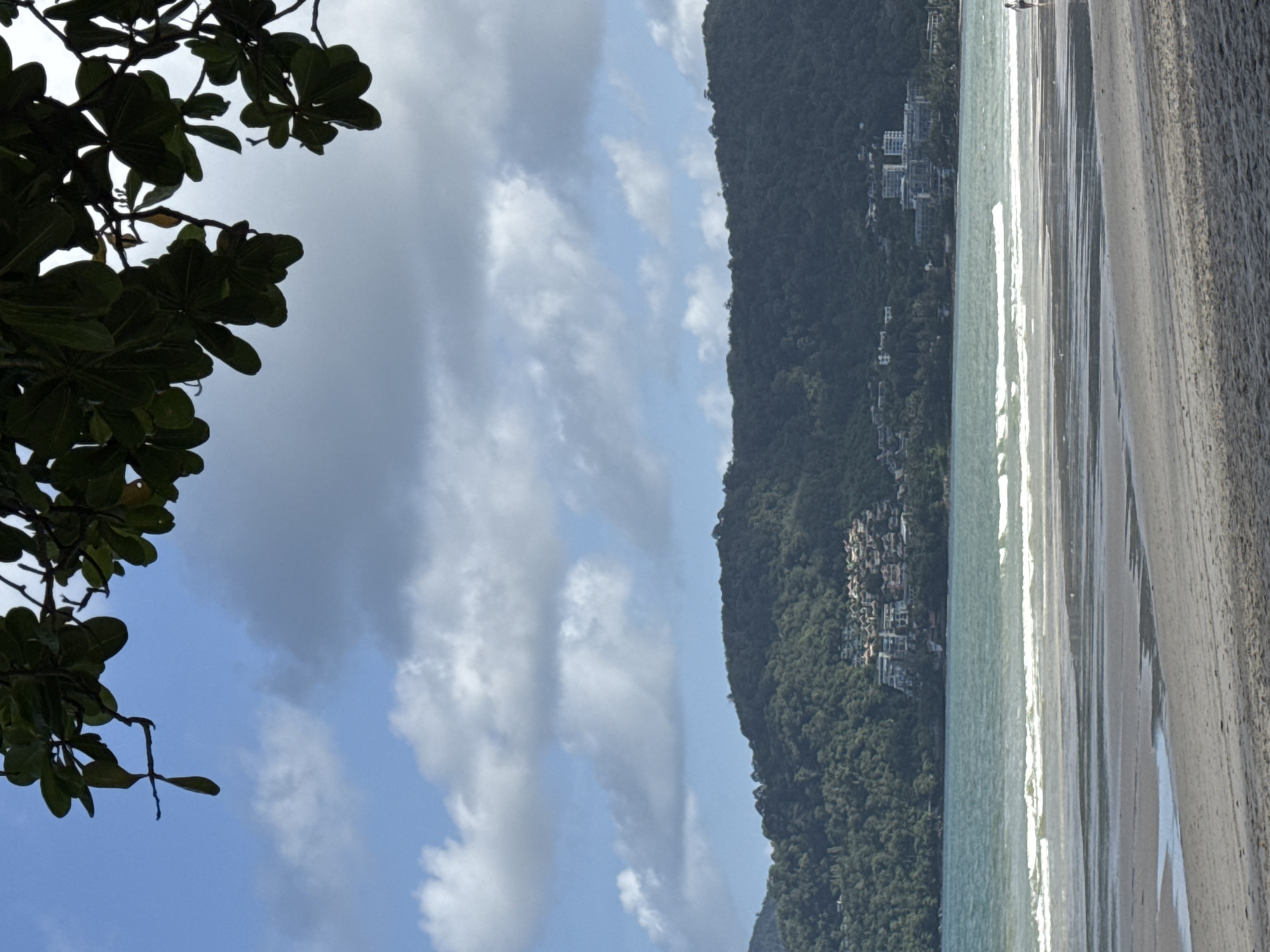 A wide shot captures a serene beach scene in Patong, Thailand. In the foreground, the sandy beach stretches out, marked by gentle tire tracks. The sea, a shimmering turquoise, laps against the shore with small, white-capped waves. In the mid-ground, a lush green mountain slopes upwards, dotted with clusters of buildings that appear to be hotels or residential complexes. The sky above is a vibrant blue, adorned with scattered, fluffy white clouds. The top of the image is framed by the dark green foliage of trees, their leaves creating a natural canopy. A lone figure, dressed in dark clothing, walks along the shoreline in the distance on the right side of the frame. The overall atmosphere is peaceful and idyllic, indicative of a tropical vacation destination.