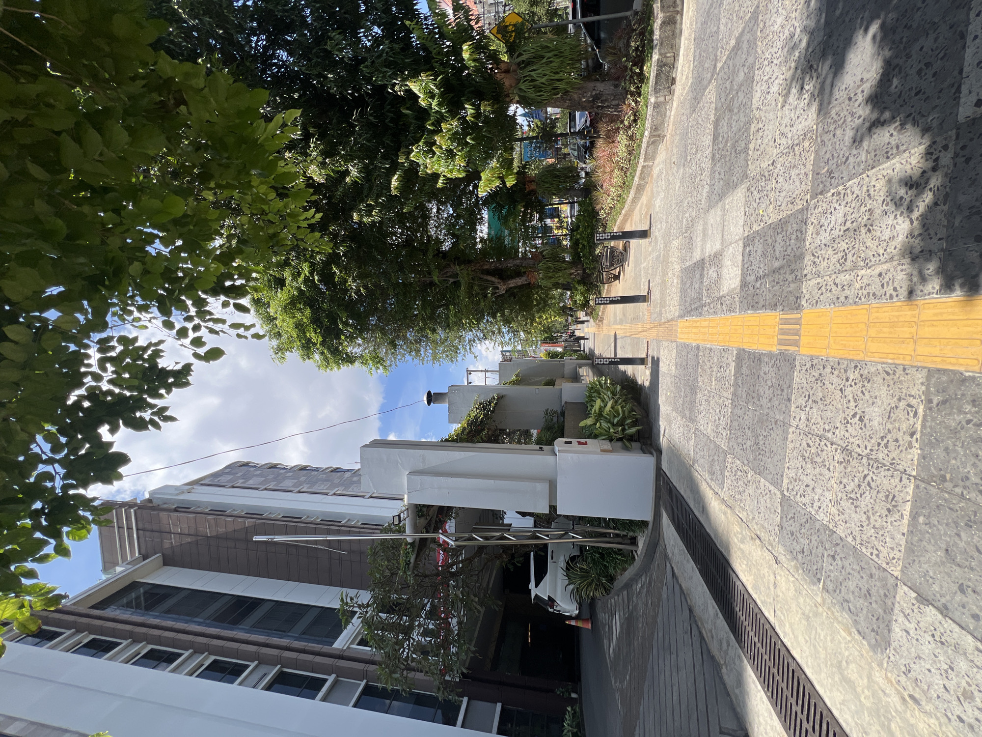 The image depicts a street scene in Yogyakarta, Indonesia. A wide paved sidewalk with textured yellow tactile paving for visually impaired individuals runs diagonally from the foreground towards the background. The sidewalk is bordered by a grey stone curb and a planter box filled with various plants, including some with large, hanging leaves. Several mature trees with dense green foliage line the sidewalk, casting shadows on the pavement.
To the left of the sidewalk, a modern building with white and brown facades rises several stories high. The building has numerous dark-tinted windows and a covered driveway or parking area is visible at the base. A white SUV is parked near the entrance of the building, and an orange traffic cone is placed on the ground.
Further down the sidewalk, there are a few people walking and some street furniture, including benches and bollards. In the background, more buildings, trees, and street signs are visible, suggesting an urban environment. The sky is bright blue with scattered white clouds, and sunlight illuminates the scene, creating distinct shadows. The overall impression is of a well-maintained urban area with a blend of modern architecture and greenery.