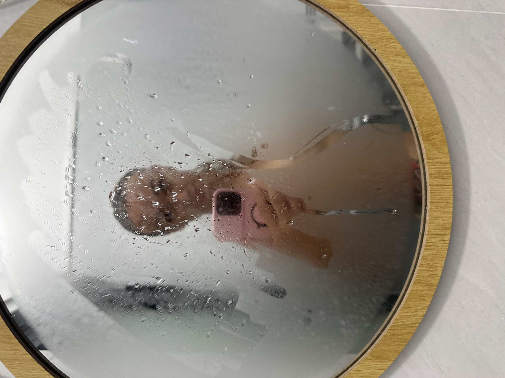 The image shows a person taking a selfie in a steamy bathroom mirror. The mirror is round and has a light wood frame. The person is holding a pink smartphone, and their face is partially obscured by water droplets and steam on the mirror. A hint of a towel rack is visible on the left side of the mirror. The background of the bathroom appears to be tiled in a light grey color. The scene is set in Mũi Né, Vietnam, suggesting it is likely a hotel bathroom given the location.