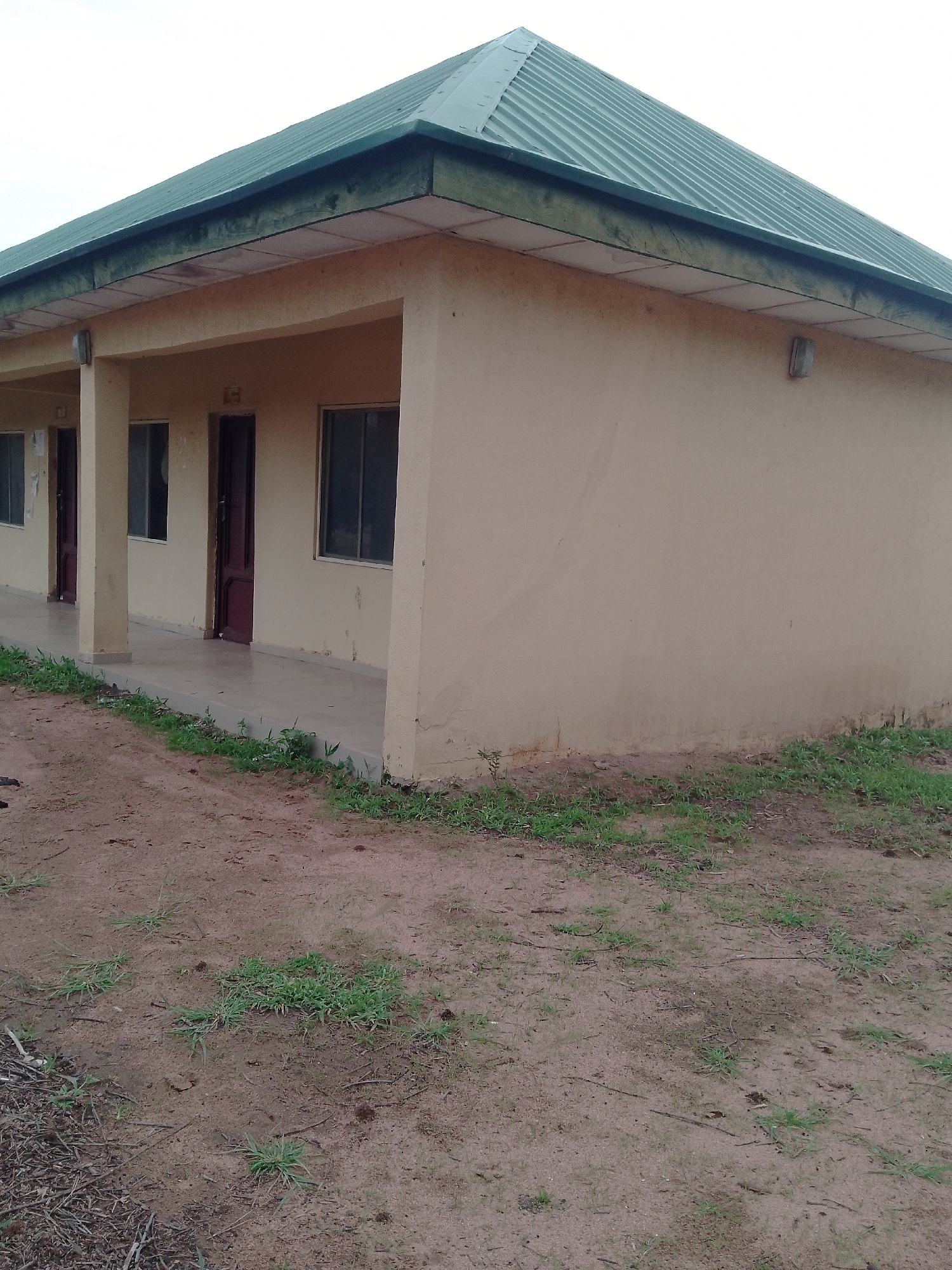 The image depicts a section of a single-story building with a green corrugated metal roof. The building has a tan-colored, stucco exterior and a covered porch area supported by rectangular pillars. Two doorways, one painted dark red and the other appearing to be open or removed, are visible along the porch. There are also two windows with dark frames. The ground in front of the building consists of sandy soil with scattered patches of dry grass and some debris. The sky above is overcast and a light gray color. The scene appears to be an outdoor shot of a building located in Jauro Yinu, Nigeria. There are no people or animals visible in the image.