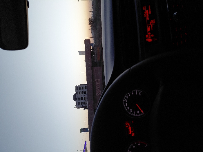A view from inside a car shows the steering wheel and dashboard, illuminated by red digital displays. Outside the window, a cityscape is visible against a pale sky, featuring buildings of varying heights, including a structure with multiple windows and another with distinctive rounded elements. A side mirror is also present in the frame.