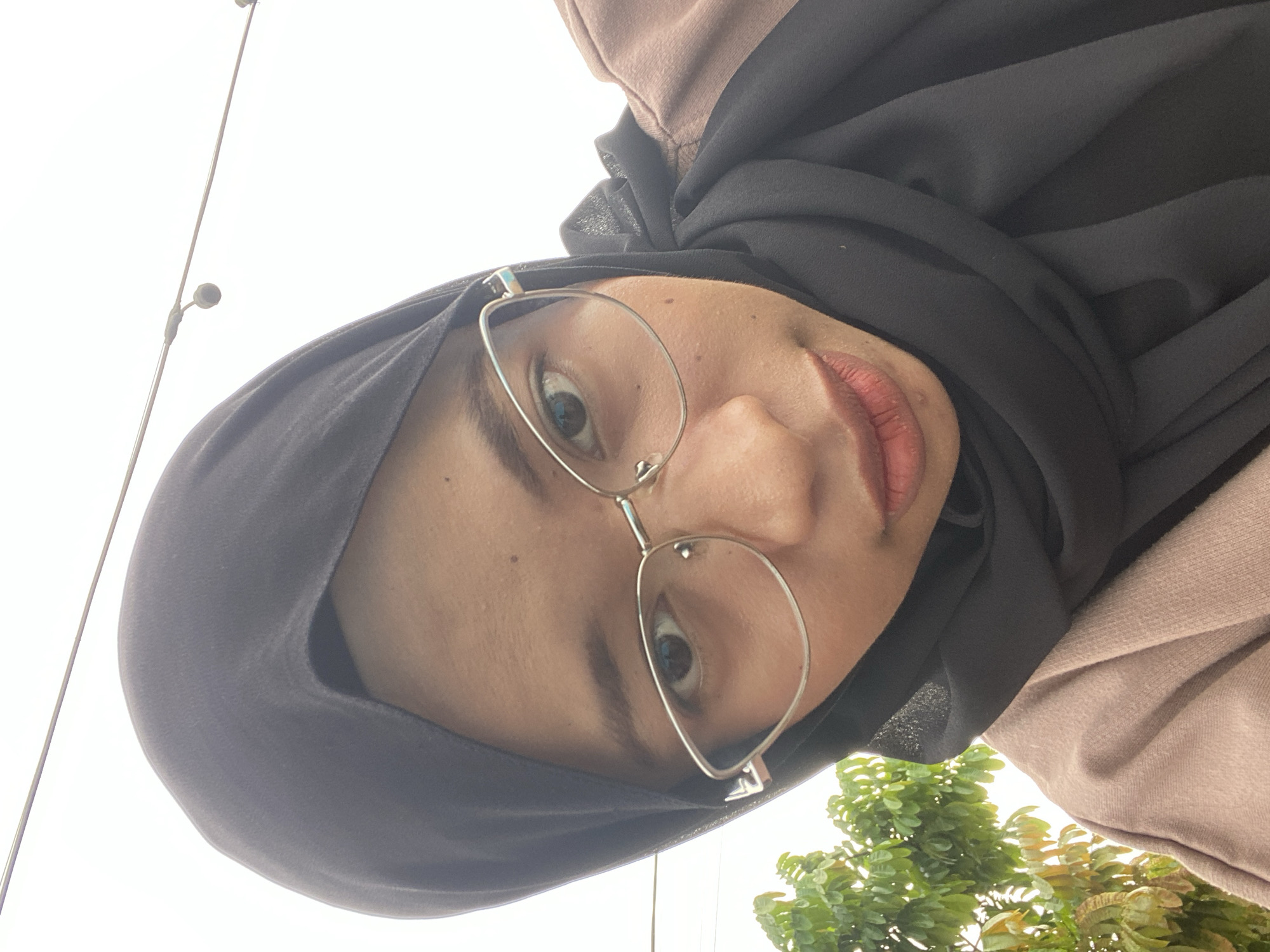 The media file shows a close-up, downward-angled selfie of a young woman wearing a black hijab and glasses. Her head is tilted, with her face filling most of the frame. She is wearing a light brown hooded top underneath the hijab. Her lips are slightly parted and appear to be painted with lipstick. The background is bright and out of focus, with a hint of green foliage on the right side, suggesting an outdoor setting. Two thin poles with small dark circular objects are visible on the left, possibly part of a street light or other outdoor fixture. The lighting suggests it is daytime, with bright, overcast conditions. The woman's expression is neutral. No text is visible in the image. The location context is Air Molek, Indonesia.