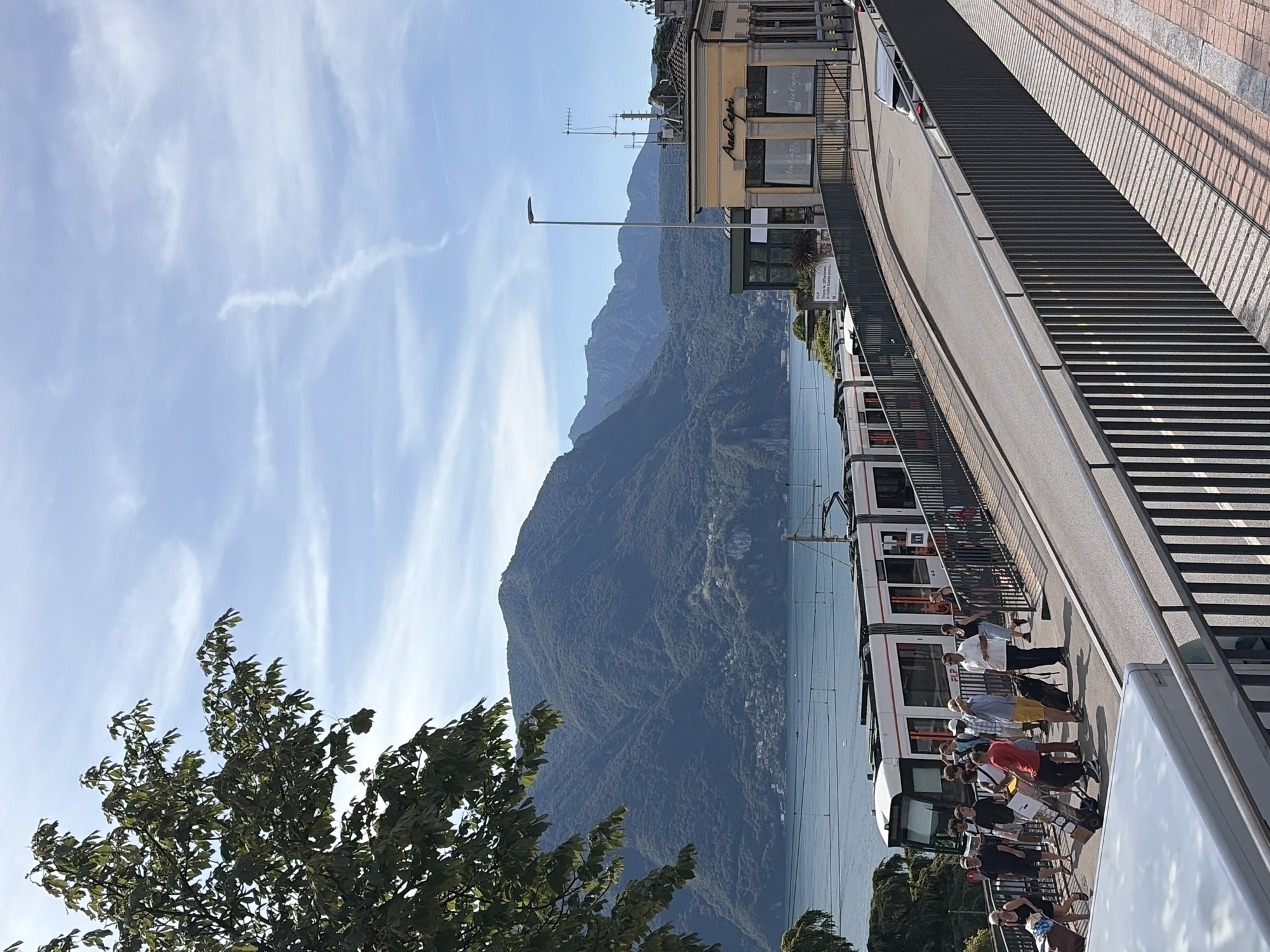 This image captures a scene along the Lugano lakefront in Lugano, Switzerland. In the foreground, a modern white tram with red accents is stopped at a station, with several passengers disembarking and waiting on a platform. People are seen with luggage, suggesting travel. To the right of the tram, a yellow building with "Ana Capri" signage appears to be a cafe or restaurant. A paved road curves upwards from the tram station area, bordered by a metal railing. Beyond the road and buildings, the expansive blue waters of Lake Lugano are visible, with lush green, tree-covered mountains rising dramatically in the background under a clear blue sky with wispy white clouds. The overall atmosphere is bright and sunny, indicative of daytime.
