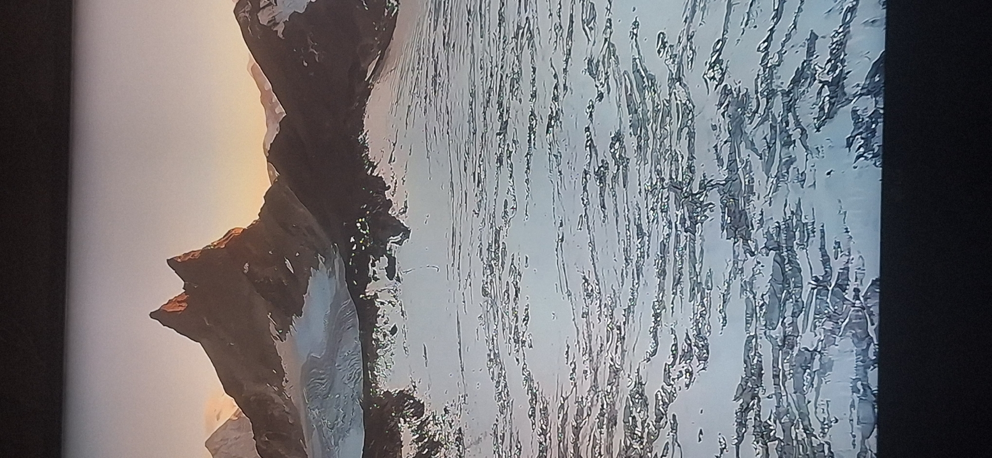The image depicts a majestic mountain landscape at dusk. Jagged, dark peaks rise sharply against a pale, orange-hued sky, their summits catching the last rays of sunlight. Snow-capped mountains stretch into the distance, with glaciers visible on their slopes. In the foreground, a vast expanse of broken ice or snow covers the ground, with textured, fractured patterns creating a sense of depth and ruggedness. The overall scene is serene and remote, evoking a sense of wild, untamed nature. There is no visible indication of the city of Bila Tserkva, Ukraine, in this image.
