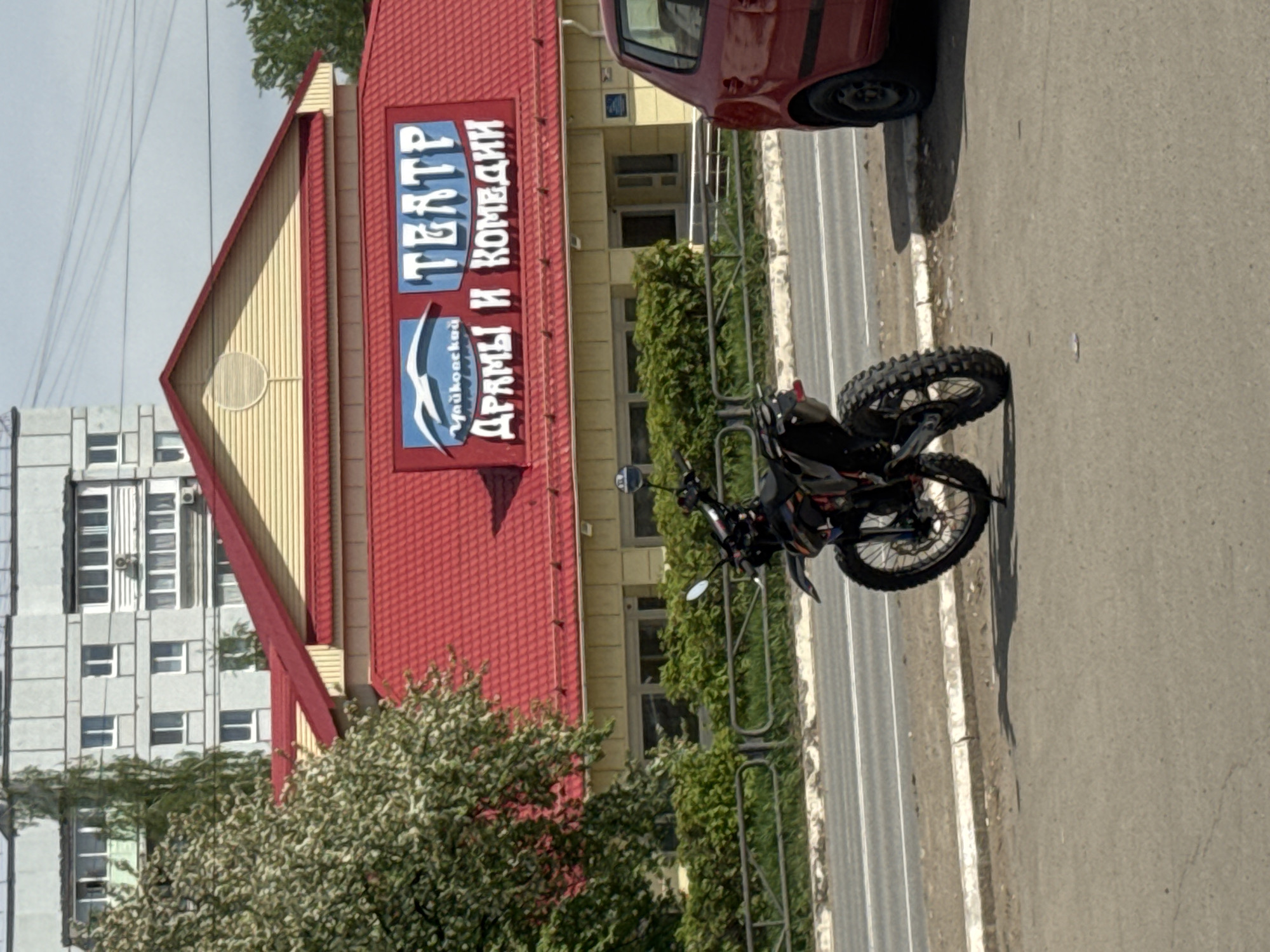 The image shows a street scene in the city of Chaikovsky, Russia. A black motorcycle is parked on the sidewalk, next to a multi-lane road. A red car is partially visible, parked behind the motorcycle. Behind the car is a yellow building with a red roof, with a sign that reads "TEATP ДРАМЫ И КОМЕДИИ Чайковского" which translates to "Drama and Comedy Theatre of Chaikovsky". A gray apartment building is visible on the left side of the frame. Greenery, including trees and bushes, is present along the sidewalk and in front of the theater. The weather appears to be sunny.