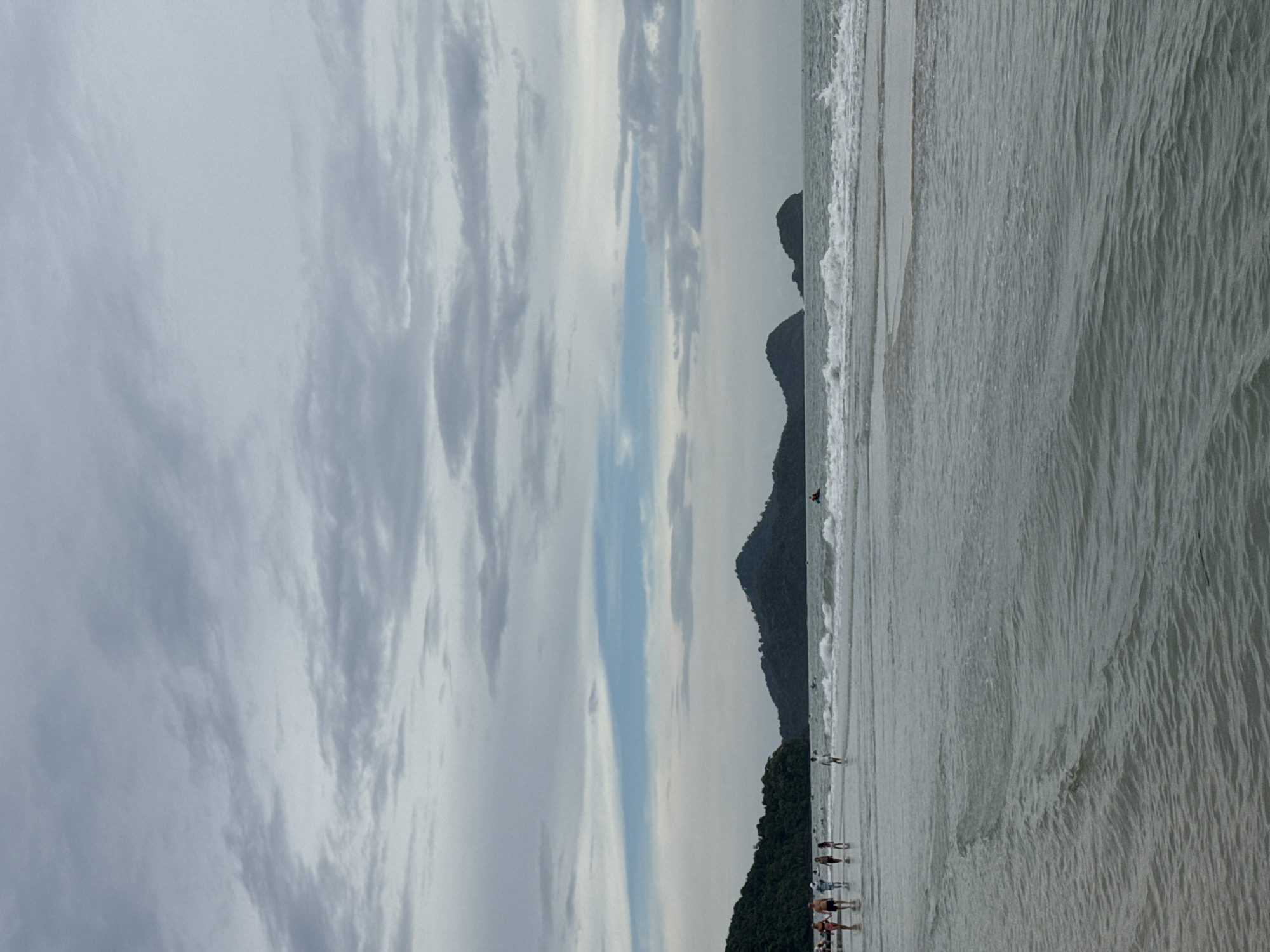The image depicts a tranquil beach scene on Tengah Island, Malaysia. The sky is overcast with grey clouds, with a faint strip of blue visible towards the horizon. The sea is calm with gentle waves lapping the shore. Several people are walking along the waterline, some in groups and some individually. In the background, lush green islands rise from the sea, creating a picturesque backdrop. The overall atmosphere is serene and peaceful, suggesting a pleasant day at the beach.
