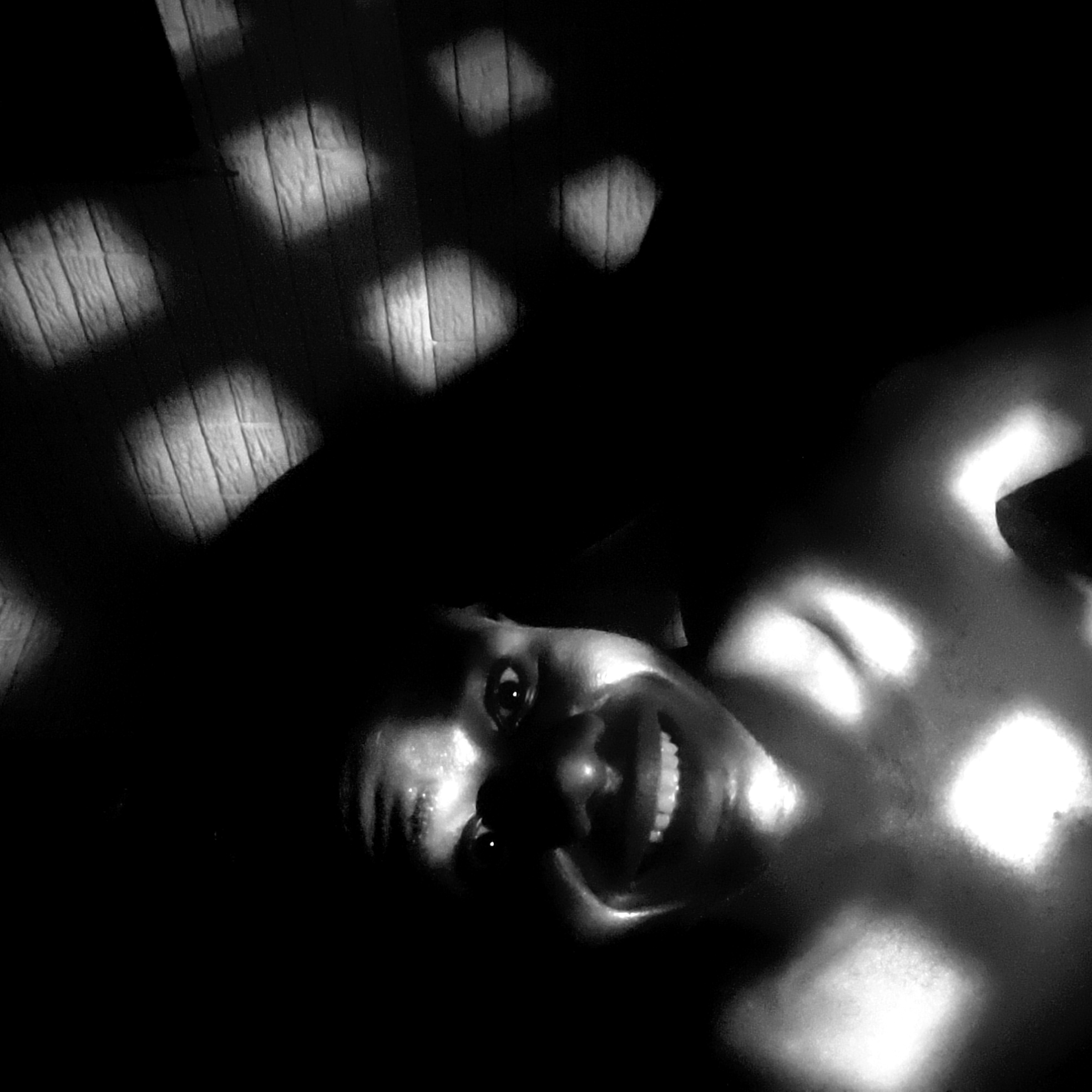 A black and white photograph features a person with dark skin, presumably an adult, lying down and facing the camera. The person is smiling broadly, revealing their teeth. The face and upper torso are illuminated by a pattern of light squares, likely caused by light filtering through a window or screen with a grid pattern. The rest of the scene is obscured by darkness. No specific details are visible that would confirm the location as Narayi, Nigeria. The photograph focuses on the subject's face and upper body with dramatic lighting.
