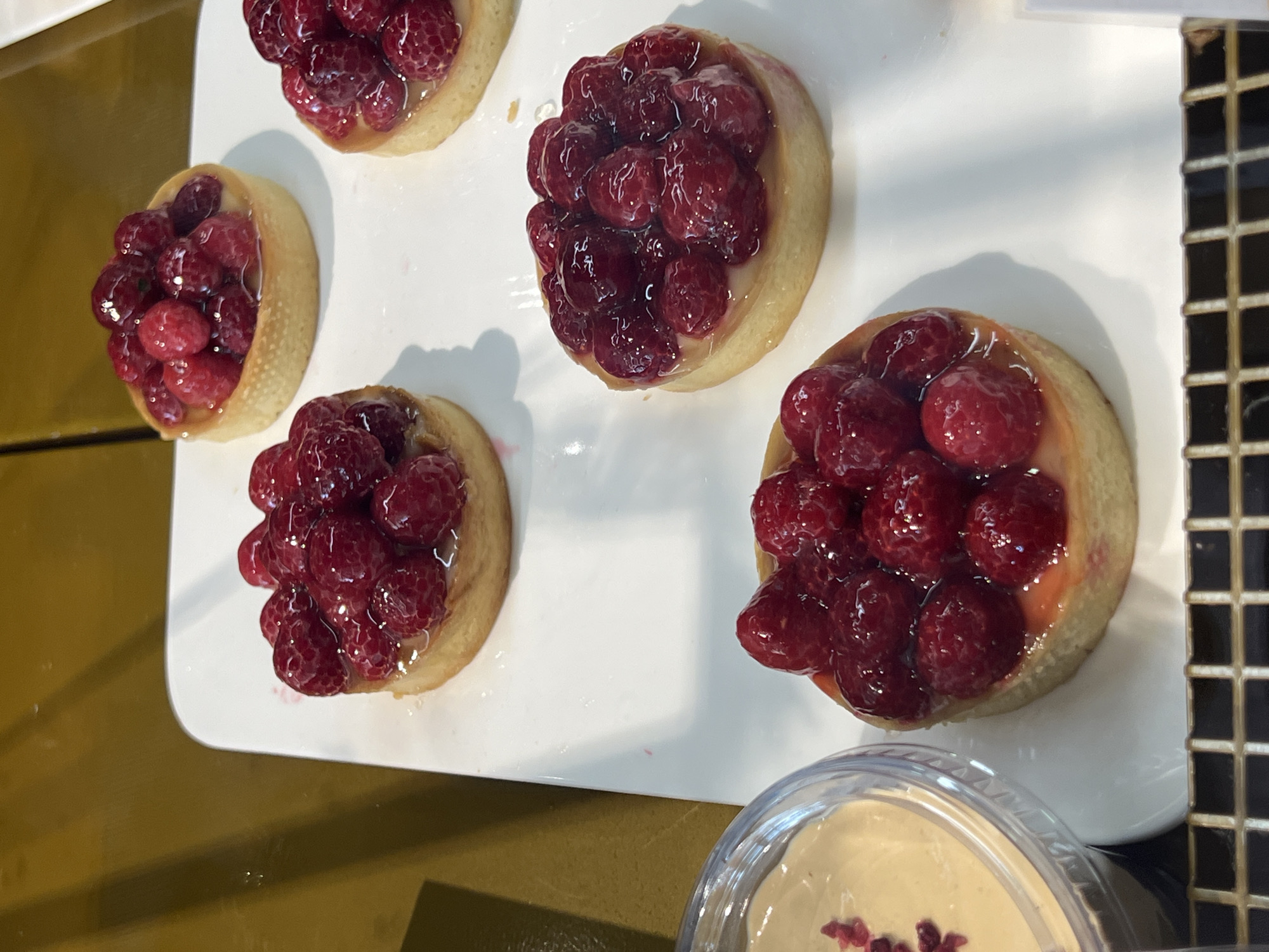 A close-up, eye-level shot shows a white rectangular platter with four raspberry tarts. The tarts are circular with a golden crust and filled with fresh raspberries. One tart is in the foreground, while the other three are arranged further back. To the left of the platter is a clear plastic container with a creamy substance and a garnish of red flakes. The background is a muted yellow-brown surface. The image is likely from a bakery or cafe.