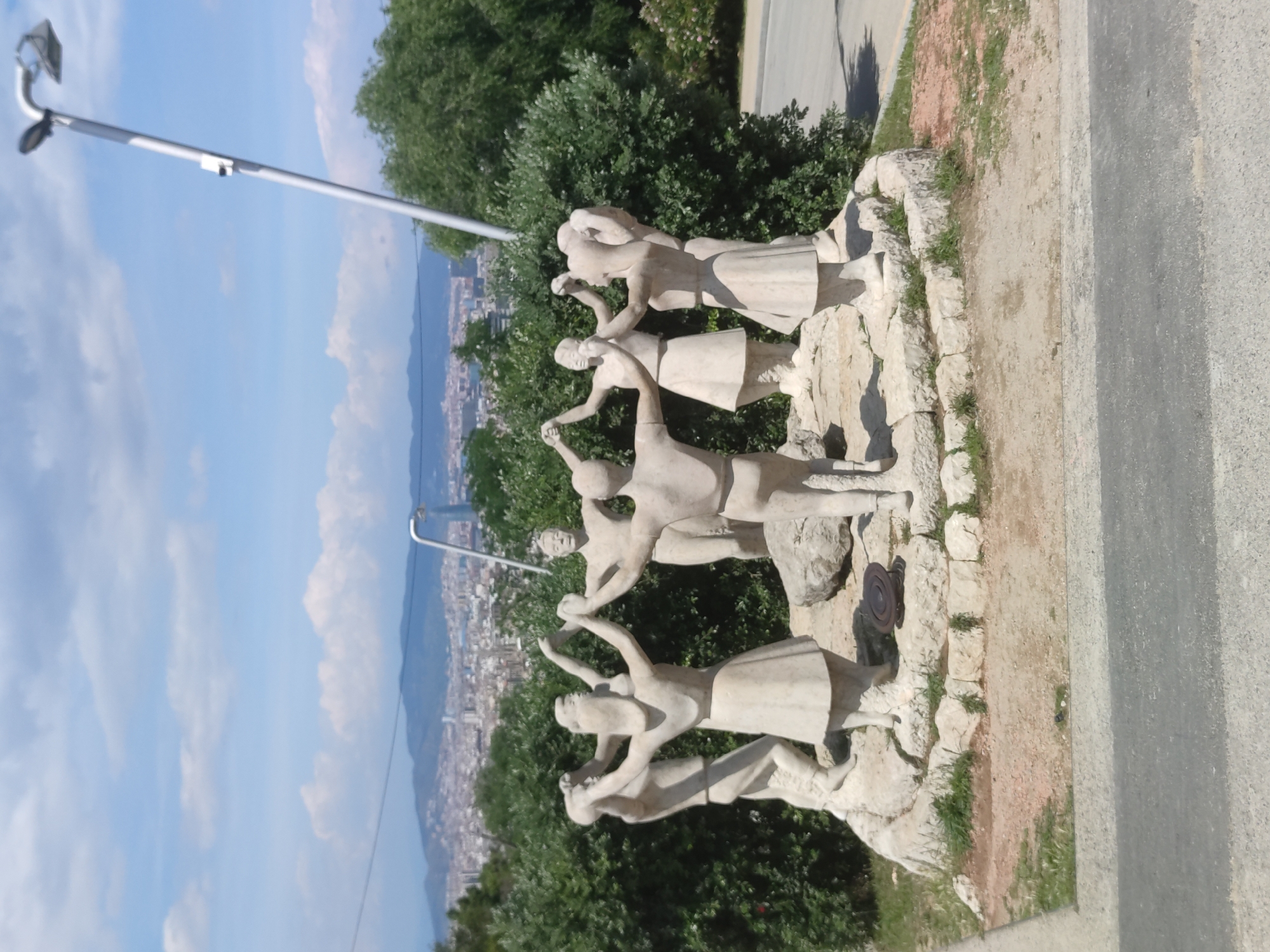 The image shows a sculpture of people dancing, likely a sardana, in Barcelona, Spain. The sculpture is positioned on a stone base, with green foliage surrounding it. In the background, there is a view of the city of Barcelona, with buildings visible and a sky with some clouds. A modern lamp post is visible on the left side of the frame. A road runs along the right side of the image.
