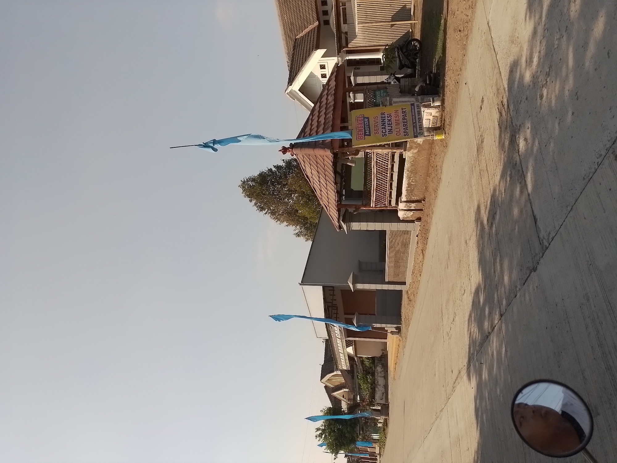 The image shows a street scene in the city of Ngancar, Indonesia. A paved road runs through the center of the frame, with a residential area to the left and right. There are several houses with tiled roofs, some with white walls and others with exposed brick. A small, open-air pavilion with a brown, pitched roof is visible on the right side of the road, in front of one of the houses. A motorcycle is parked to the right of the pavilion. A blue flag is flying from a pole in front of the pavilion, and another blue flag is visible further down the street to the left. A sign with Indonesian text advertising "BENGKEL MOTOR" (motorcycle workshop) and services such as "SERVIS," "SCANNER INJEKSI," and "OLI MESIN SPAREPART" is placed near the pavilion. The sky is clear and pale blue, suggesting daytime. In the foreground, a reflection of a person's face is visible in a motorcycle's rearview mirror. The overall scene depicts a quiet street with signs of local commerce and residential life.