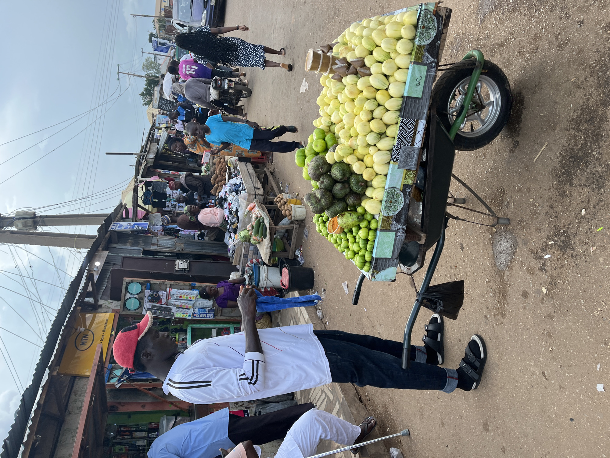 The image captures a street scene in Gura Top, Nigeria, with a vendor standing next to a cart laden with fruits. The vendor, a man wearing a red cap and a white shirt with black stripes, is looking down at his hands, which are holding a blue cloth. He is dressed in dark jeans and black sandals. The cart, a green wheelbarrow, is piled high with a variety of fruits. Prominently displayed are light yellow, oval-shaped fruits, possibly melons or cantaloupes, and a darker green, bumpy fruit, which could be avocados or a similar type of produce. Some smaller green fruits, possibly limes or small tomatoes, are also visible.
Behind the vendor, stalls and shops line the street, constructed from corrugated metal and wood. A prominent yellow sign with the "MTN" logo and text indicating "Data Broadband" is visible on one of the structures. People are seen browsing or walking through the market area. In the background, a blue shirted young boy walks past, and further down the street, a woman with long dark hair in a patterned dress walks away from the camera. The ground is paved with reddish-brown dirt and gravel, with some scattered debris. The sky is overcast, suggesting cloudy weather. Electrical wires are strung across the top of the scene, supported by utility poles. The overall impression is of a bustling, open-air market environment.