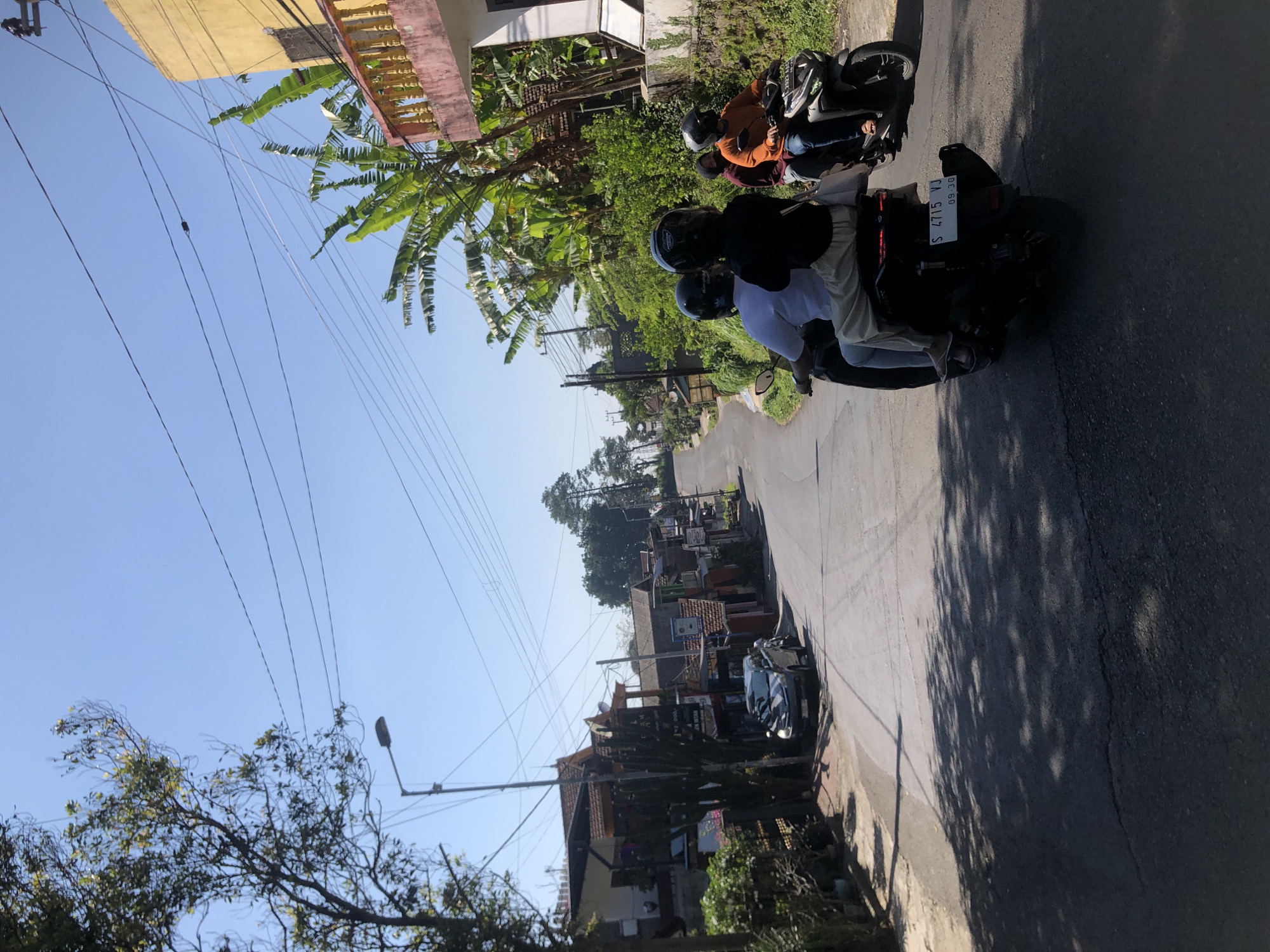 The image depicts a sunny day in Trawas, Indonesia, on a paved, uphill road lined with buildings and lush vegetation. The perspective is from a low angle, looking up the road. Two motorcycles are traveling uphill. The first motorcycle, closer to the viewer and in the foreground, carries three people: a driver wearing a white shirt and black helmet, a passenger behind him in a black shirt and helmet, and a person in a black shirt and helmet behind the passenger. The second motorcycle, further up the road, carries two people: a driver in an orange shirt and helmet and a passenger in a dark top and mask, also wearing a helmet. Both motorcycles have license plates.
On the left side of the road, several buildings with tiled roofs are visible, some appearing to be shops or residences, with trees and plants in front of them. A black car is parked on the left side of the road. On the right side, there is a yellow building with a balcony and pink railings, and more dense greenery, including banana trees. Overhead, power lines are strung across the clear blue sky. The road surface shows some wear and tear, with shadows of trees cast across it, indicating a bright, sunny environment.