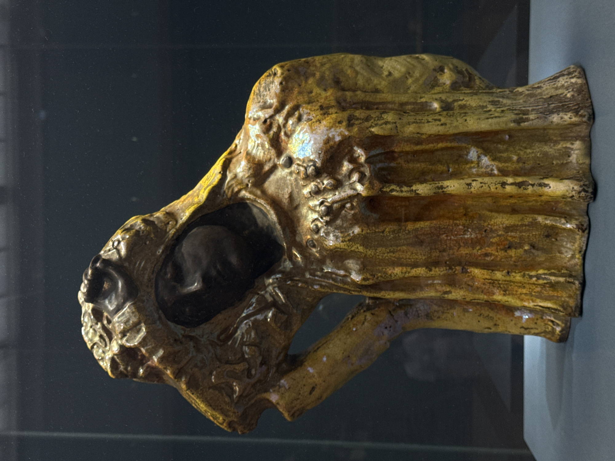 A close-up, side view shows an aged, yellow-brown, sculpted artifact with textured surfaces. A dark figure is set within an opening near the left end. The object rests on a gray ledge, likely within a display case in a museum. The setting is likely the Pushkin State Museum of Fine Arts in Moscow, Russia, which houses similar antiquities.
