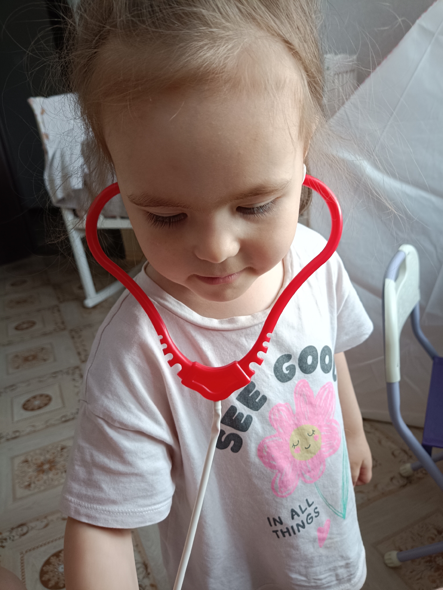 The image shows a close-up, high-angle portrait of a young child, likely a girl, engaged in imaginative play indoors in Noviy, Russia. The child has light blonde or light brown hair pulled back, and her face is focused downwards, with her eyes cast down, suggesting concentration on an activity. She is wearing a light-colored, short-sleeved T-shirt.
A prominent red plastic toy stethoscope is draped around her neck, with the earpieces resting in her ears, indicating she is playing doctor. The T-shirt features a graphic design: a large pink flower with a yellow center and a smiling face, surrounded by green leaves. Below the flower, text is visible that reads, "SEE GOOD" (the "D" and part of "IN ALL THINGS" with a small pink heart below it.
The background reveals an indoor setting. A patterned floor, possibly tiled in shades of brown and beige, is visible on the left. On the right side, parts of light-colored furniture with purple accents are discernible, along with a glimpse of a white fabric or curtain. The lighting suggests it is daytime. The child's absorbed expression conveys an innocent and playful mood.