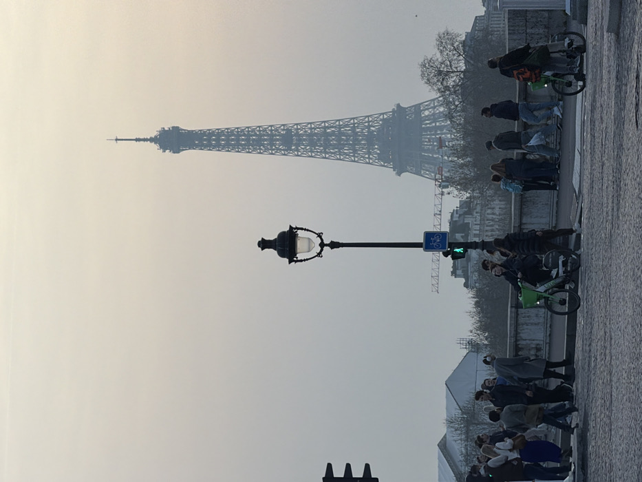 The image displays a hazy, overcast scene in Paris, France, focused on a street-level view with the Eiffel Tower prominently positioned in the background. The tower's intricate lattice structure is visible through a diffused atmospheric haze.
In the mid-ground, a traditional black Parisian streetlamp stands upright, featuring a spherical luminaire. Below the lamp, a blue circular sign with a white bicycle symbol indicates a bicycle route, and a green pedestrian signal is illuminated further down the pole. To the far left, a section of a black traffic light array is visible. The immediate foreground shows cobblestone pavement.
Approximately 15-20 individuals are present on a pedestrian area or bridge. On the left, a group of people walks, including one individual in a long grey coat with headphones. To the right of the streetlamp, a person sits on a bright green Lime share-bike, facing left. Further to the right, another individual on a similar green Lime bike faces away from the viewer, carrying an orange bag on their back. Other pedestrians are observed standing or walking along a light-colored stone parapet that delineates the mid-ground.
Behind the parapet and a sparse line of leafless trees, indistinct Parisian buildings are visible, typical of Haussmannian architecture. A red construction crane is visible above these buildings, situated to the left of the Eiffel Tower's base. The overall lighting is soft and uniform, suggesting an early morning or late afternoon with low visibility.