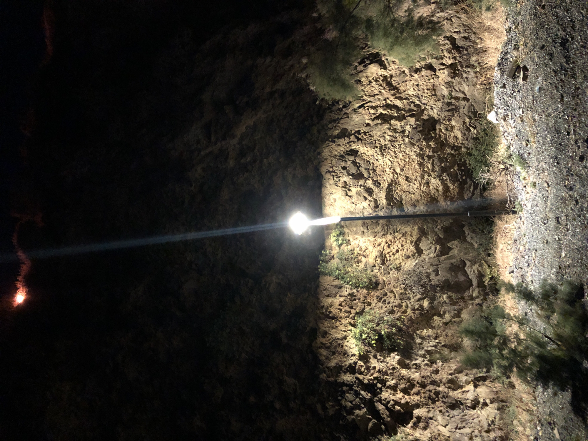 The image shows a street light illuminating a rocky hillside at night. The hillside is covered in rough, uneven stones and sparse vegetation, including small bushes and dry grass. A single street light, emitting a bright white light, is positioned in the center of the frame, casting a strong beam upwards and illuminating the rock face. The ground in the foreground is covered in gravel and debris. The sky is dark, with a faint reddish glow visible at the top of the image, possibly from a distant light source or fire. The scene appears to be located in a semi-rural or undeveloped area, possibly on the outskirts of a town. The location is identified as Karaburun, Turkey.