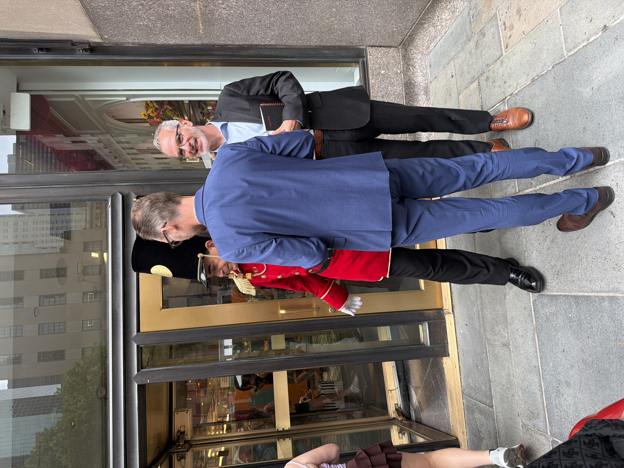 In front of a building with large glass doors, a person in a red uniform with a tall black hat stands guard. To the right, two men in suits converse. The man on the left wears a blue suit, and the man on the right wears a dark suit and glasses, holding a black notebook. The setting appears to be an outdoor urban environment, likely a city street with buildings reflected in the glass doors, suggesting a location in New York City, United States of America. The ground is paved with rectangular stones.