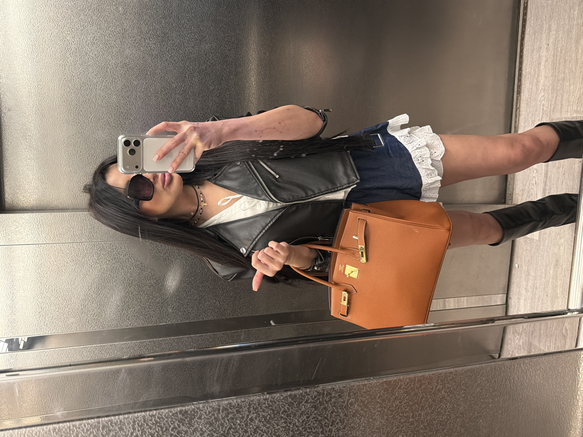 A woman is taking a selfie in an elevator. She is wearing sunglasses, a black leather jacket, a white top, a blue skirt with lace trim, and black boots. She is holding a tan-colored handbag. The elevator walls are metal with a textured finish.