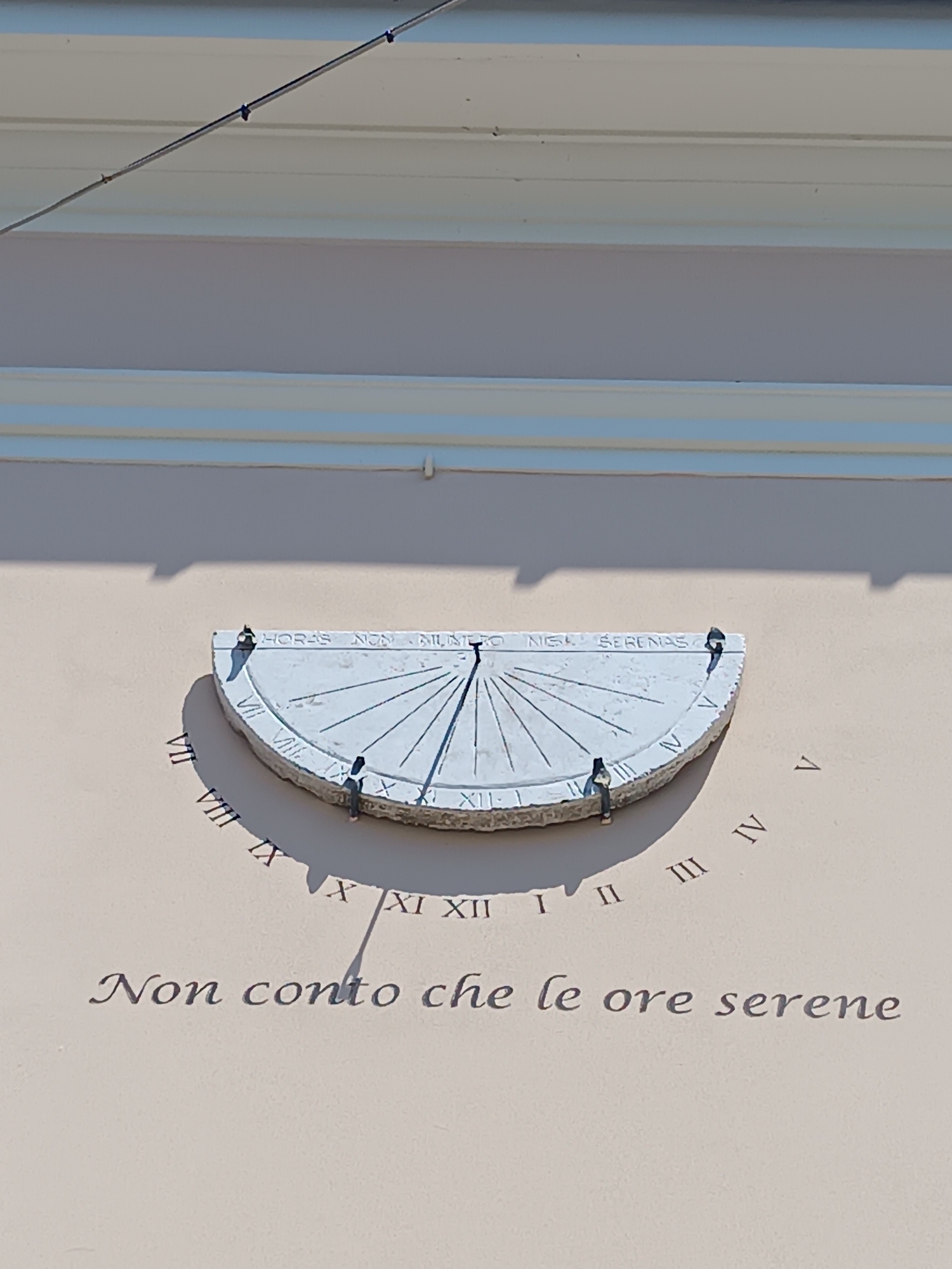 The image shows a sundial mounted on a light pink wall, likely part of a building in Mezzana Sant'Antonio, Italy. The sundial is a semi-circular stone structure with Roman numerals marking the hours. The gnomon casts a shadow indicating the approximate time. The Latin phrase "HORAS NON NUMERO NISI SERENAS" is inscribed on the dial. Below the sundial, the Italian phrase "Non conto che le ore serene" is written on the wall. A white trim runs along the top of the wall. A thin electrical wire runs above the trim.
