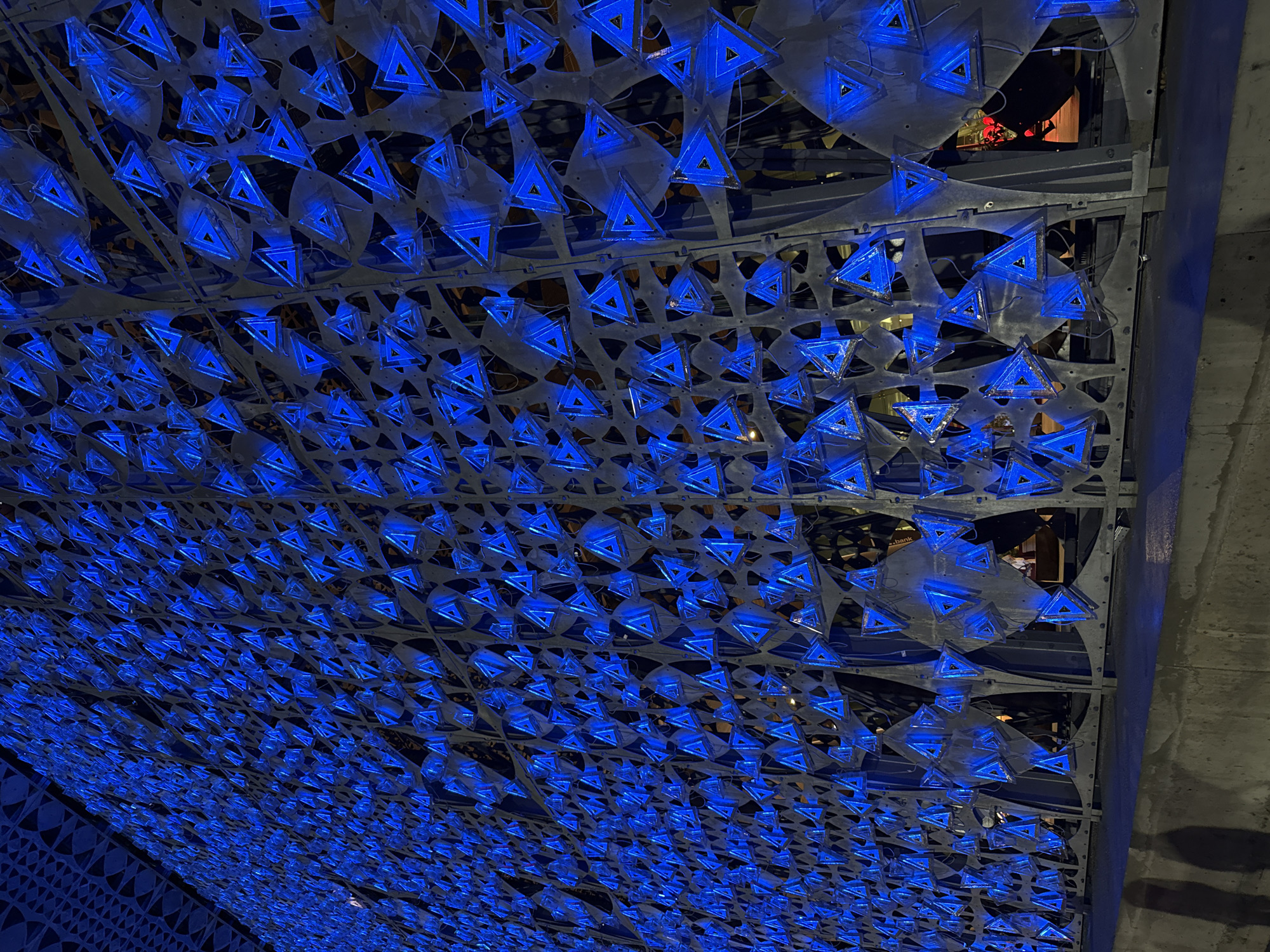 The image depicts an illuminated, triangular mesh structure on the exterior of a building in the city of Kruja, Albania. The structure is a large, dark metal grate with countless small, hollow triangles embedded within it. These triangles are illuminated from within by a bright, electric blue light, creating a vibrant and intricate pattern. The lighting casts a blue glow on the surrounding area and reflects off the metallic surfaces of the structure. In the foreground, a concrete ledge or sidewalk is visible, with the shadows of two people cast upon it. The background is dark and somewhat obscured, suggesting it is nighttime. The overall scene is modern and artistic, with the blue lights creating a striking visual effect against the dark backdrop.