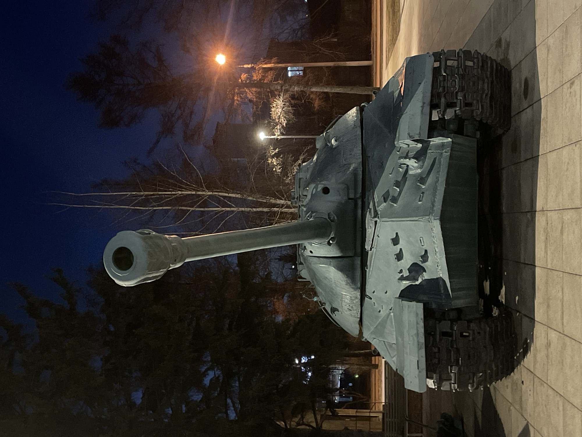 The image depicts a Soviet-era tank monument, likely a T-34 or similar model, situated in an outdoor setting. The tank is painted in a muted blue-gray color. Damage or wear and tear can be seen on the tank's armor. The background is dark, suggesting it is dusk or night, with illuminated streetlights visible. Bare trees stand behind the tank, with buildings partially visible. The tank is positioned on a paved surface, likely a sidewalk or plaza. The exact location is unknown.