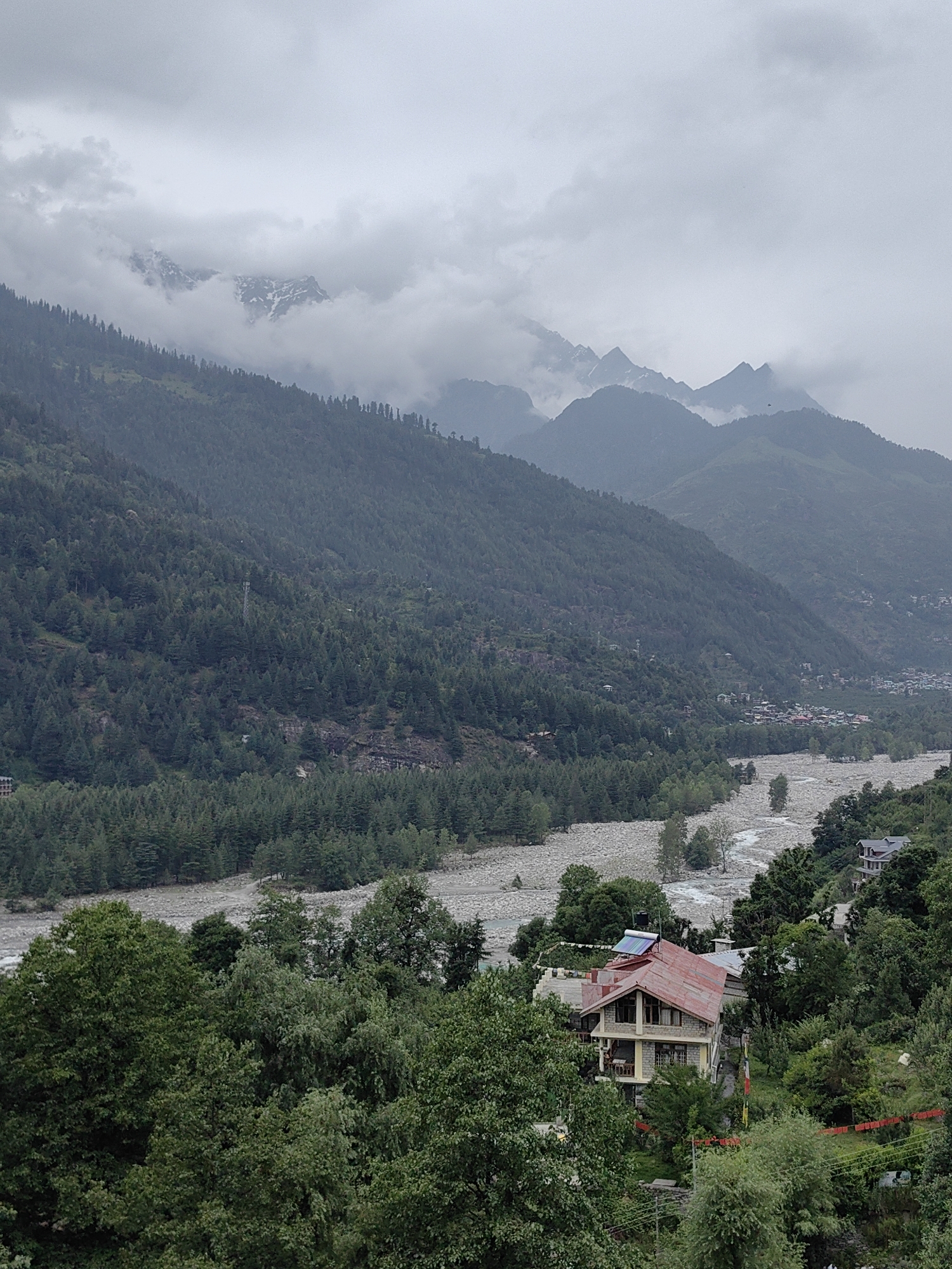 The image depicts a mountain landscape in Vashisht, India, characterized by a cloudy sky and lush green vegetation. In the foreground, a cluster of trees frames a multi-story building with a red roof, possibly a guesthouse or hotel, surrounded by more foliage. The middle ground features a winding riverbed filled with pebbles, bordered by a dense forest of pine trees that extend up the slopes of the mountains. In the background, rugged mountain peaks are visible, partially obscured by clouds and mist, with remnants of snow on some of the higher elevations. The overall atmosphere is serene and natural, with hints of human settlement integrated into the environment.