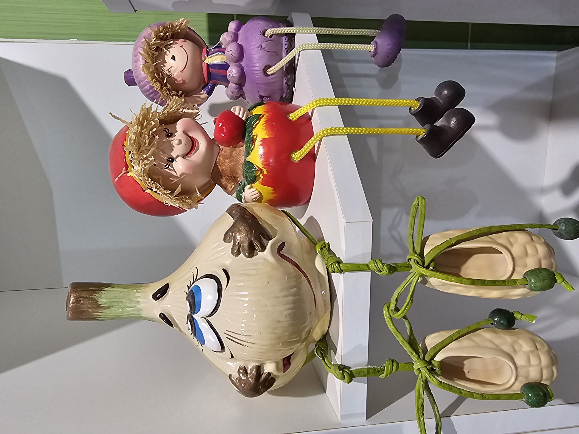 This image depicts a still life arrangement on a white shelf. The primary subjects are three decorative figures: a large, tan ceramic garlic head with blue eyes and brown hands resting on its cheeks, a girl figure with a red hat and straw hair sitting on a red and yellow apple-shaped base, and a second, smaller girl figure with a purple hat and striped dress sitting on a purple base. Both girl figures have their legs hanging down, with the first girl's legs attached to brown boots and the second girl's legs attached to purple shoes. Hanging below the garlic figure are two tan ceramic shoe-like objects, each with green twine and green bead accents. The background consists of a white wall and a hint of green vertical surface on the right. The lighting appears to be artificial. This scene is located in the city of Kalinovka, Russia.
