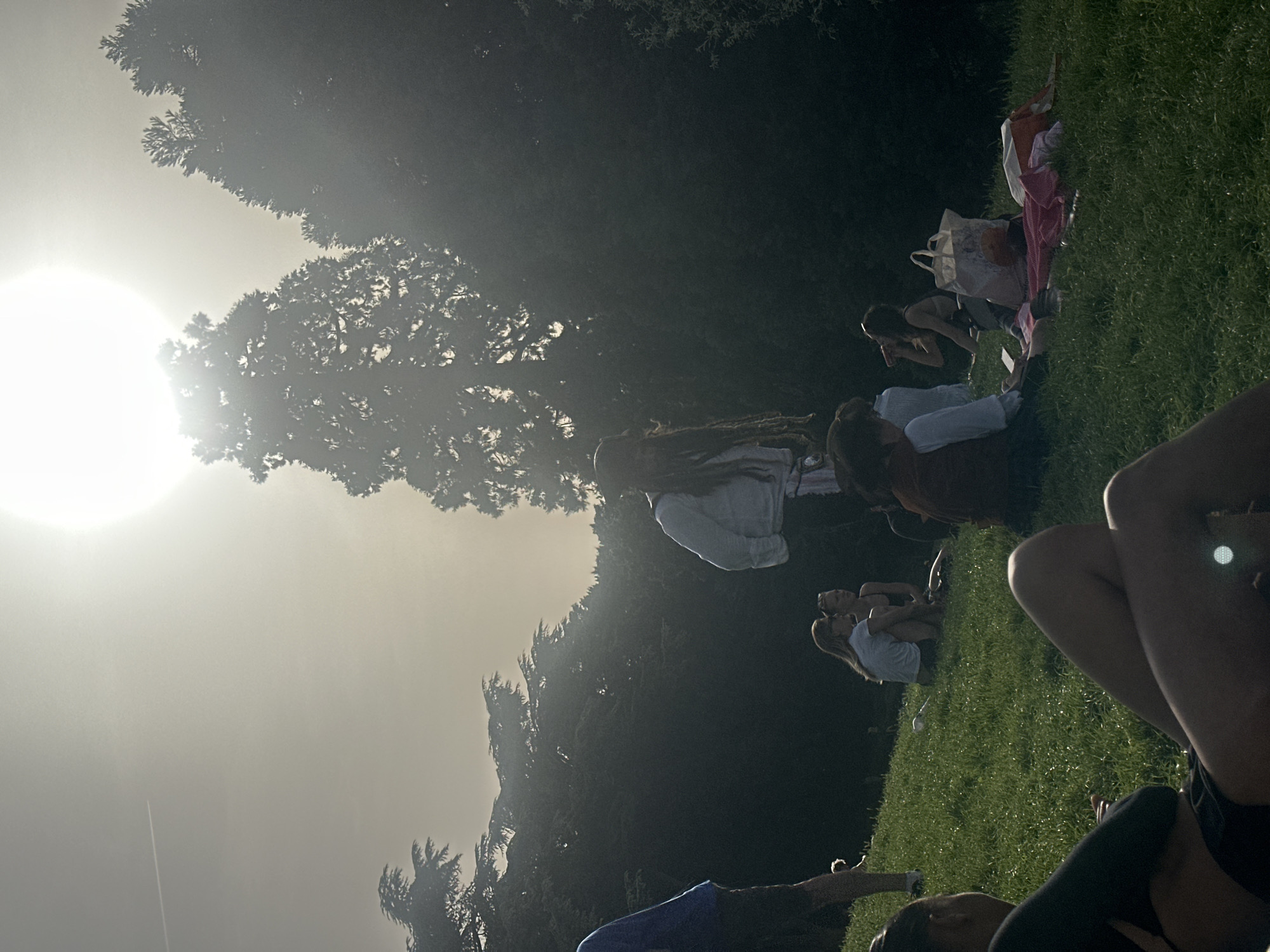 The image shows a group of people relaxing on a grassy hill in what appears to be a park on a sunny day. The sun is bright, obscuring some details due to lens flare. Some people are sitting, lying down, or interacting in small groups. One person is kneeling and looking into a bag on the ground. There are blankets and bags scattered around on the grass. Trees are in the background. It is likely this is in Paris, France.