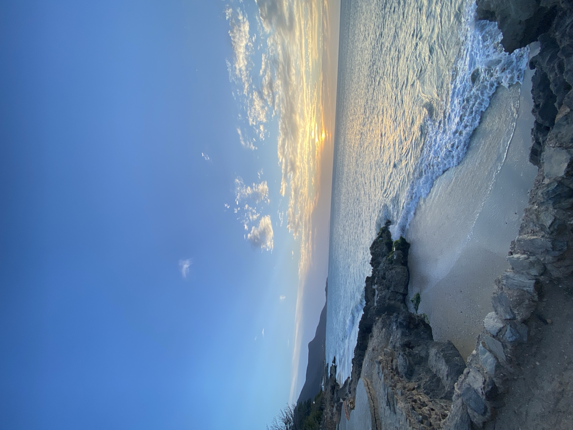 This image captures a coastal scene likely in Juan Griego, Venezuela, suggested by the landscape and seascape details. The photograph is taken from an elevated position, possibly a rocky outcrop or cliffside path, looking towards the ocean. The sun appears to be setting or rising, casting a golden glow on the water and illuminating the underside of scattered clouds in the blue sky. Waves are breaking along a sandy beach adjacent to the rocky terrain. There is a mountain or hill visible in the distance near the shoreline. The foreground includes rocky formations and a paved path, suggesting a developed coastal area. No people are directly visible in the frame.