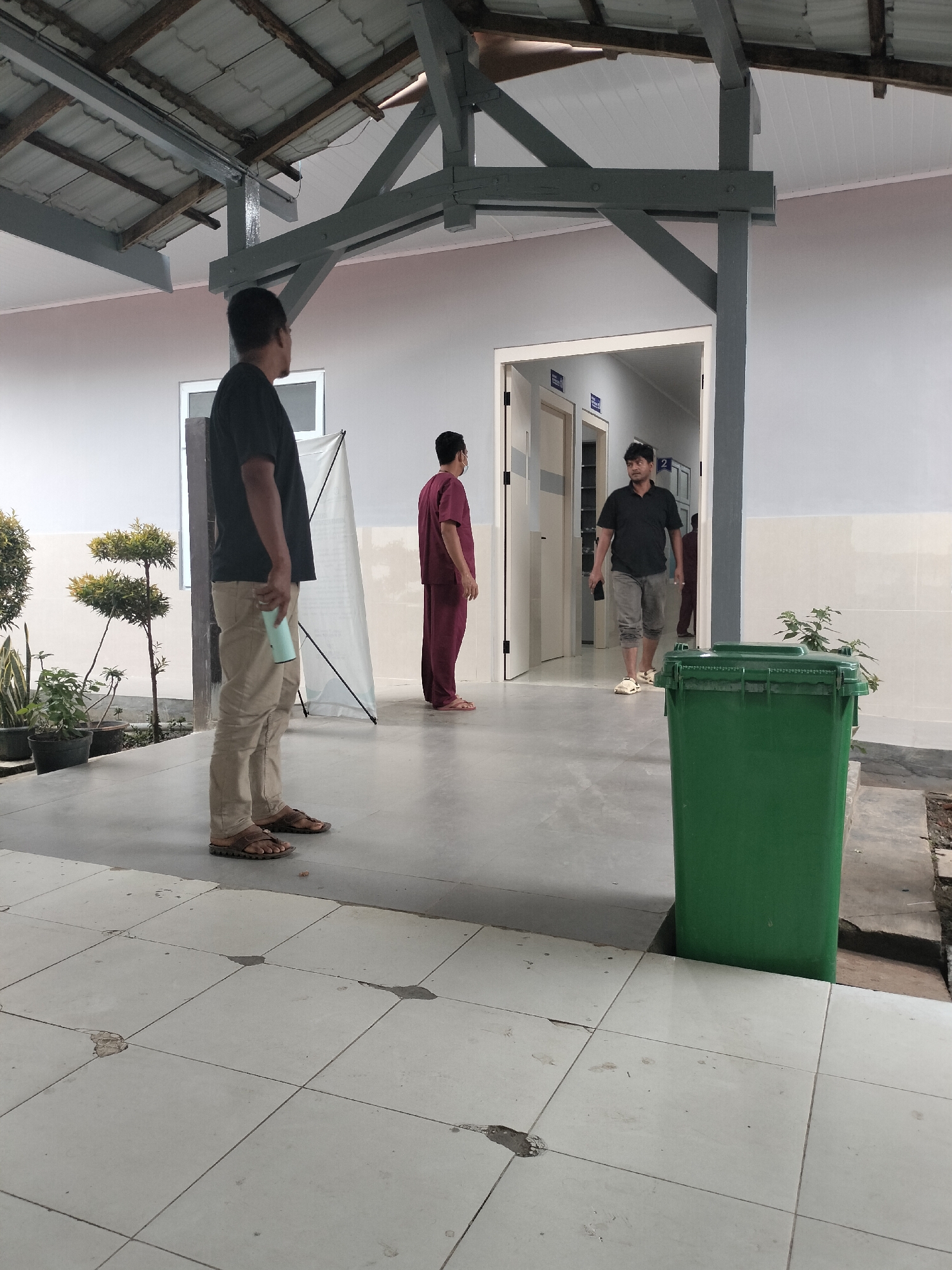 The image depicts an outdoor covered walkway, likely part of a medical facility in Langsa, Indonesia, given the context of the image. Three men are visible. In the foreground on the left, a man wearing a black t-shirt and tan pants stands facing away from the camera, holding a light blue object. To his right, further into the scene, a man in a maroon uniform stands facing towards an open doorway. Inside the doorway, a third man in a black t-shirt and grey shorts walks away from the camera. A large green trash bin is positioned on the right side of the walkway. The walkway is paved with light-colored tiles, some of which show signs of wear. The roof of the walkway is supported by visible grey beams. Potted plants are situated on the left side of the walkway. The overall scene appears to be an ordinary day at a public or medical institution.