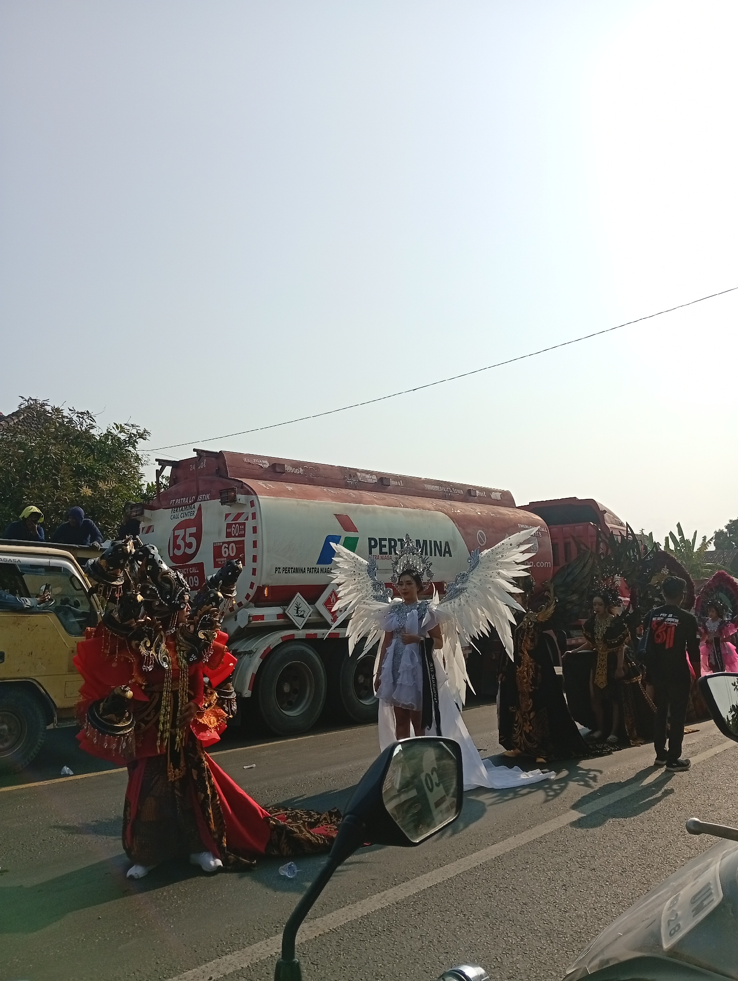 In the city of Sarang, Indonesia, a street parade is taking place, featuring individuals in elaborate costumes. In the foreground, a person adorned in a striking red and gold traditional outfit with intricate headdress and a long train is positioned on the left side of the road. To their right, a person wearing a white costume with large, feathery wings and a crown stands prominently. Behind them, several other individuals are dressed in dark, ornate costumes, and a few more figures in colorful attire can be seen further down the road.
A large Pertamina fuel tanker truck, identifiable by its branding and white and red livery, is parked on the road, serving as a backdrop to the parade participants. A yellow truck is also visible on the left, with several people standing on top and behind it. The scene unfolds on a paved road under a clear, bright sky, with trees and buildings visible in the background, suggesting an outdoor urban or semi-urban setting. The overall atmosphere appears festive, characteristic of a parade or cultural event.