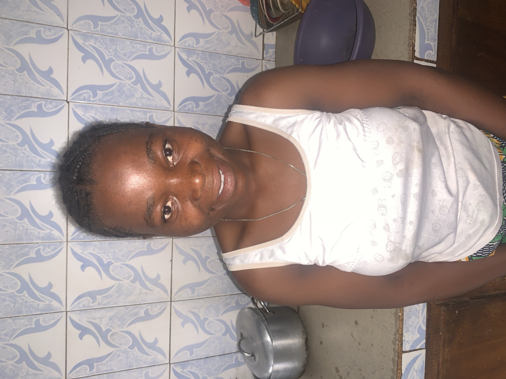 A woman stands in a room with patterned blue and white tiles on the wall. She is wearing a white tank top and a silver necklace. A metal cooking pot sits on the floor near her. A purple bowl and drying rack with dishes are visible in the background. The location is likely a kitchen, potentially in a home or apartment in Jos, Nigeria, but definitive confirmation of the city is impossible without additional information.