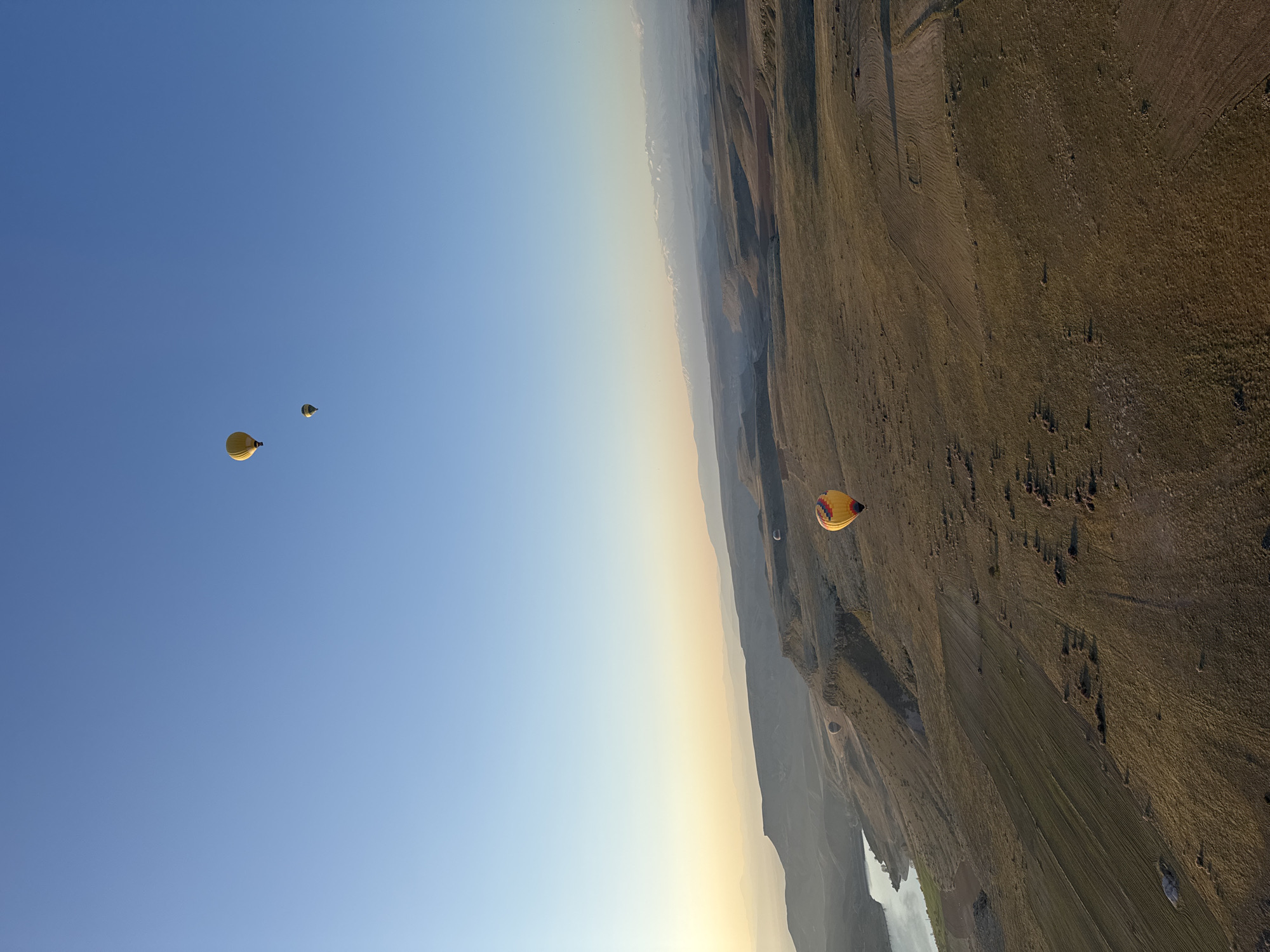This aerial photograph captures a serene hot air balloon ride over the vast, rugged landscape of Yeşilhisar, Turkey, likely during sunrise or sunset. The scene is presented from a tilted perspective, emphasizing the expansive sky and the undulating terrain below.
The sky, occupying the upper portion of the image, transitions from a deep, clear blue overhead to a soft, hazy yellow and orange glow near the horizon, indicative of either early morning or late afternoon light. Several other hot air balloons dot the sky and landscape. One prominent yellow balloon is visible in the upper left, and another, featuring yellow, red, and blue panels, is seen closer to the center-right, seemingly lower and nearer to the ground. A smaller, darker balloon is visible further in the distance.
The landscape below consists of rolling hills and valleys covered in dry, golden-brown grass and agricultural fields. Patches of tilled land with distinct patterns are visible, alongside areas of more natural scrub. A winding body of water, possibly a river or a lake, can be seen snaking through the distant valley. The soft, warm light casts long shadows across the terrain, highlighting its contours and giving depth to the scene. The distant horizon is characterized by a soft atmospheric haze, suggesting mountains or further plains fading into the distance. No visible text is present in the image.