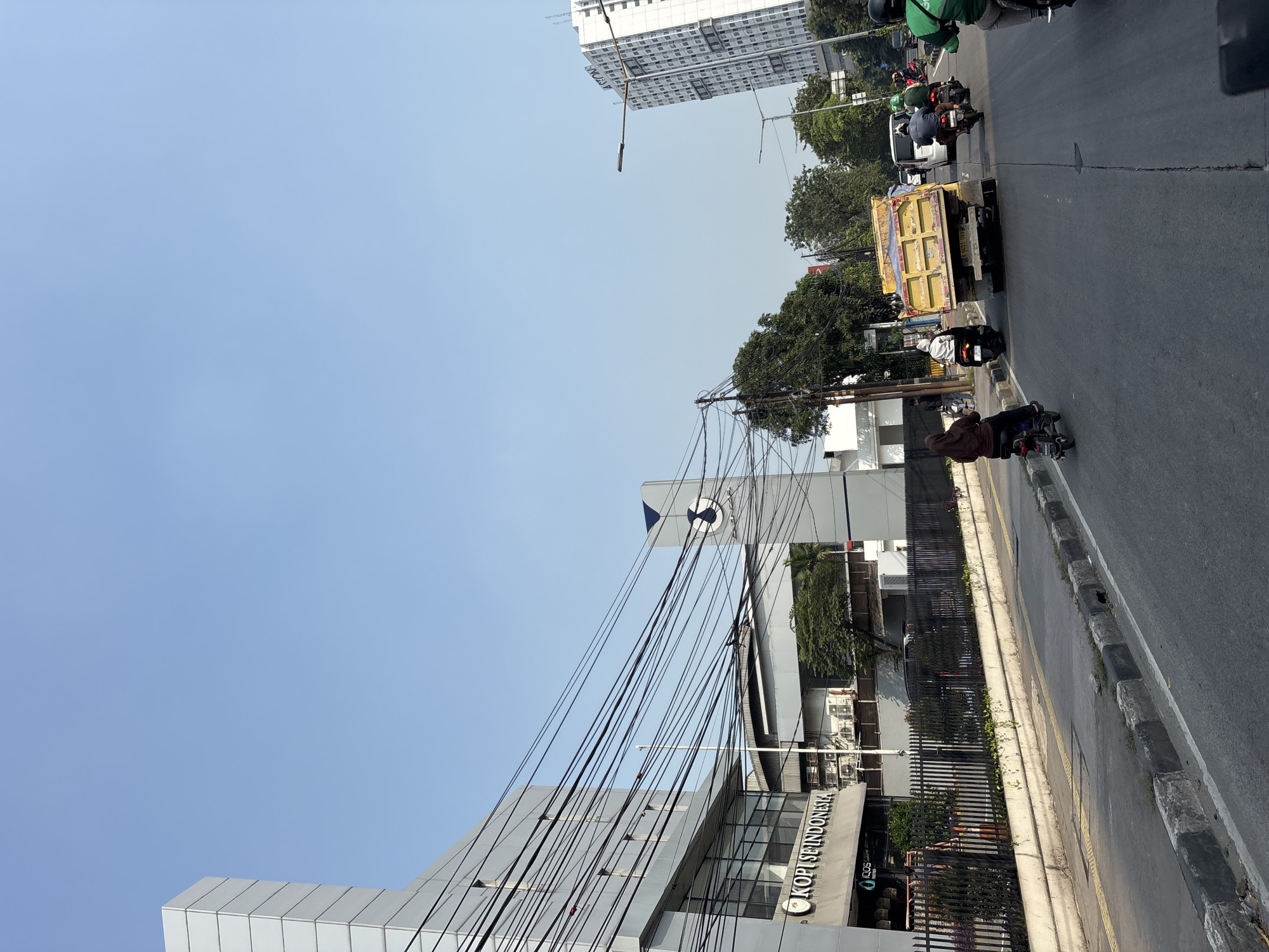The image depicts a street scene in North Jakarta, Indonesia, on a clear day. A modern, multi-story building with a gray facade and numerous horizontal windows dominates the left side of the frame. Prominently displayed on the building is a sign reading "KOPI SE INDONESIA". A complex network of dark electrical wires crisscrosses the upper portion of the image, strung between poles and the building.
In the middle ground, a yellow dump truck is the most prominent vehicle on the road, with its bed raised. Several motorcycles are visible, including one rider in a brown headscarf on the left, and others behind the dump truck, some with passengers. A white van is also present in the traffic. To the right, a tall, white apartment building with many windows is visible in the distance. Green trees line the sides of the road in the background, and more vegetation is seen behind a black metal fence in front of the "KOPI SE INDONESIA" building. The road is paved, with a distinct curb and sidewalk. The sky is a clear, light blue.