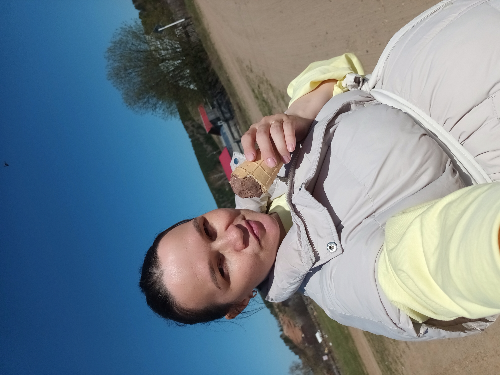 A woman is holding a partially eaten chocolate ice cream cone. She is wearing a light yellow shirt and a light gray jacket. She has a ring on her finger. The background shows a dirt road, a building with a red roof, trees, and a clear blue sky. The location is possibly Ольховка, Russia.
