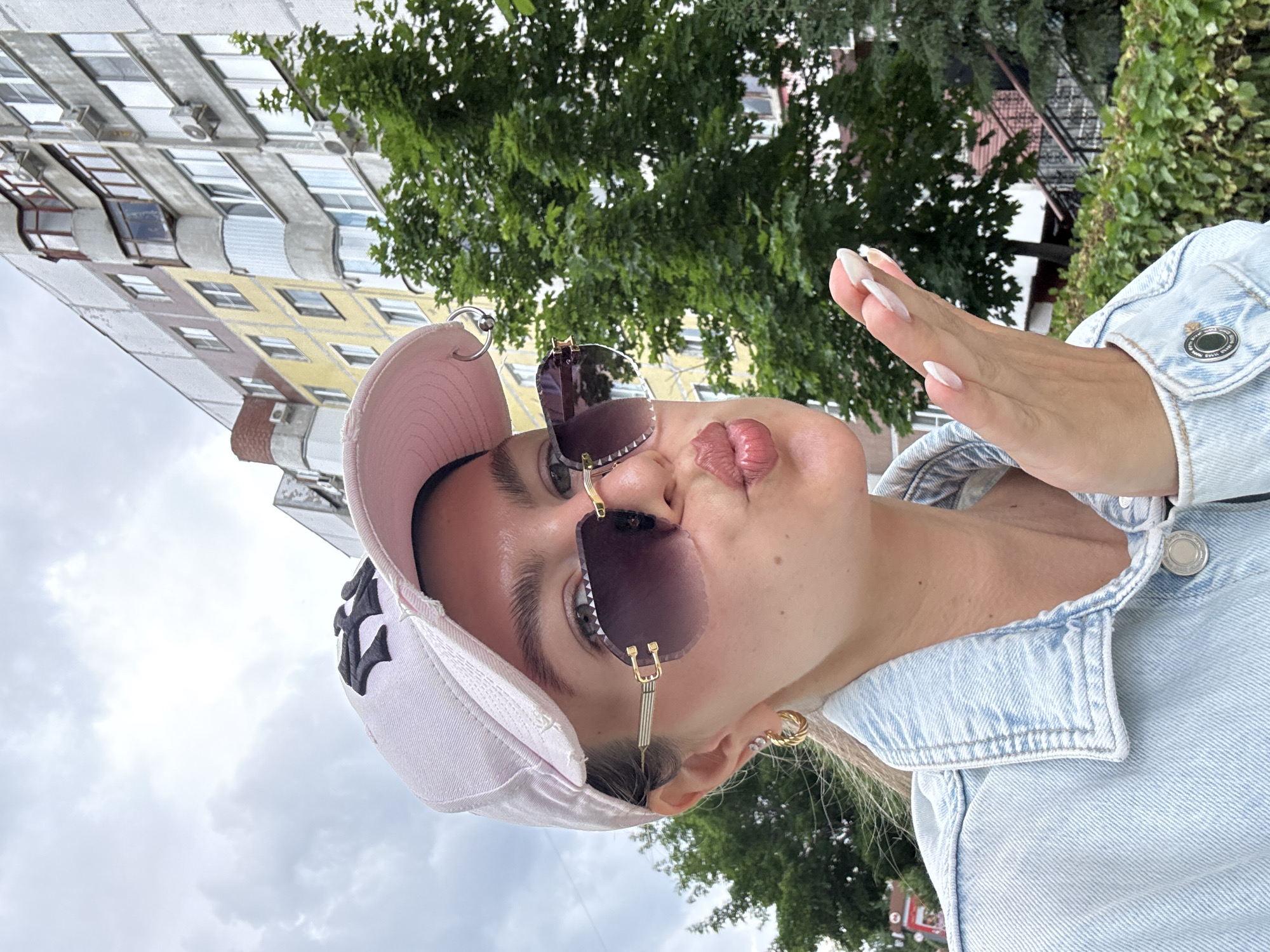 In this medium-shot, eye-level outdoor selfie, a young white woman is depicted from the chest up, facing the camera with a pout and blowing a kiss. She is wearing a light blue denim jacket over a light-colored top, a pink baseball cap with a black logo, and rimless sunglasses with gold accents. Her blonde hair is pulled back, and she has noticeable makeup, including defined eyebrows and lipstick. She is wearing gold hoop earrings and has long, manicured fingernails. Her right hand is extended towards the camera, fingers spread. The background features a multi-story apartment building with a yellow and grey facade, several air conditioning units, and lush green trees and bushes. The sky is overcast with clouds. The location is identified as Unknown, Unknown.