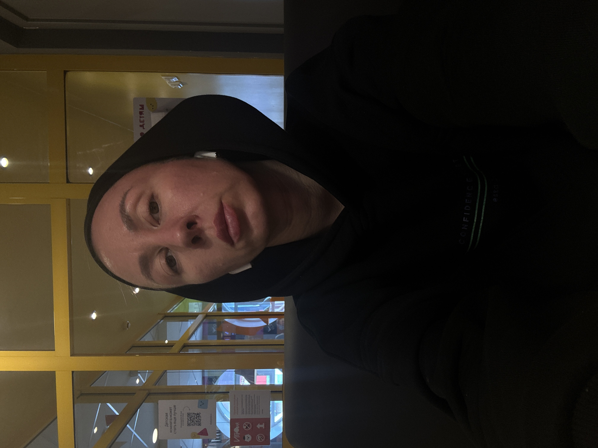 The image captures a person, likely an adult, in an indoor setting. The individual is wearing a black hooded sweatshirt with the hood up, and white wireless earbuds are visible in their ears. They are looking towards the camera with a neutral expression, lips slightly parted.
The background features glass partitions framed by distinctive bright yellow struts, suggesting a commercial or public building environment such as a mall, cafe, or waiting area. Overhead spotlights are reflected in the glass. The person appears to be seated on a dark, upholstered surface, possibly a bench or sofa.
Notable details include Cyrillic text visible in the background, consistent with the Votkinsk, Russia context. On the person's sweatshirt, the word "CONFIDENCE" is printed vertically in a light color. In the background, reflected on the glass, the word "ДЕТЯМ" (FOR CHILDREN) can be seen. Another sign, partially visible, reads "ДЕТСКАЯ комната может стать еще лучше" (CHILDREN'S room can become even better) and includes QR codes and small illustrative icons. The lighting suggests an indoor scene, potentially during daytime given the ambient light coming through the background. The overall impression is one of a relaxed moment in a public interior space.