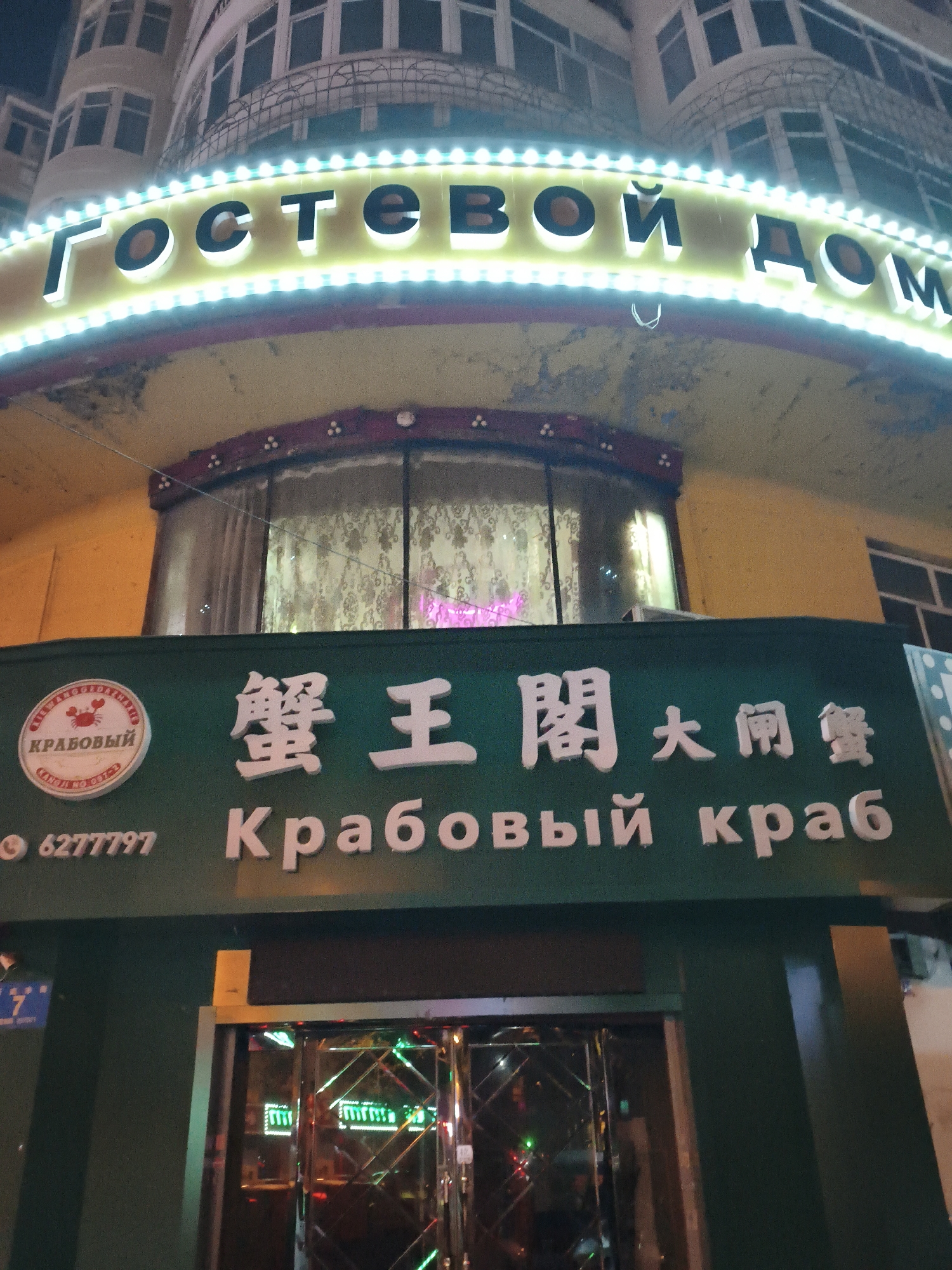 An exterior view at night captures a brightly lit building with signage. The prominent yellow sign at the top features illuminated Cyrillic text that reads "Гостевой Дом" (Guest House). Below this, a dark green sign displays a circular emblem with a crab and the Cyrillic text "КРАБОВЫЙ" (Crab). To the right of the emblem, larger white characters in what appears to be Chinese are visible, followed by more Cyrillic text: "Крабовый краб" (Crab crab). A phone number "62777797" is also present on the green sign. The building itself appears to be multi-storied with arched windows. The entrance area is visible at the bottom of the frame, featuring reflective doors and interior lighting. The overall scene suggests a commercial establishment, possibly a restaurant or inn, in Xi'an, China, given the mixture of Cyrillic and Chinese text.