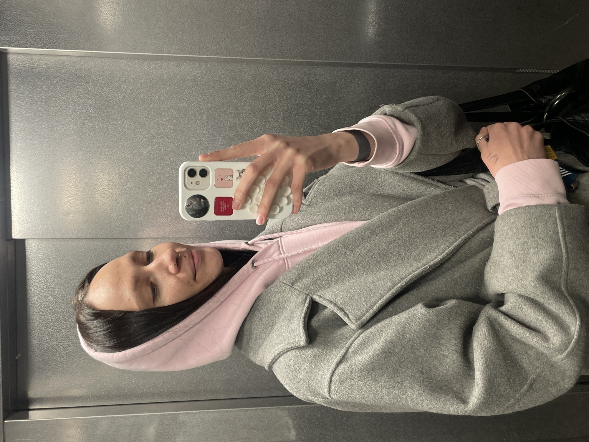 A woman is taking a selfie in what appears to be an elevator. She is wearing a pink hoodie and a gray coat. Her phone has a white case with stickers on it. The background consists of the interior of the elevator, featuring a metallic wall. There is no specific information to suggest the location is in Izhevsk, Russia.