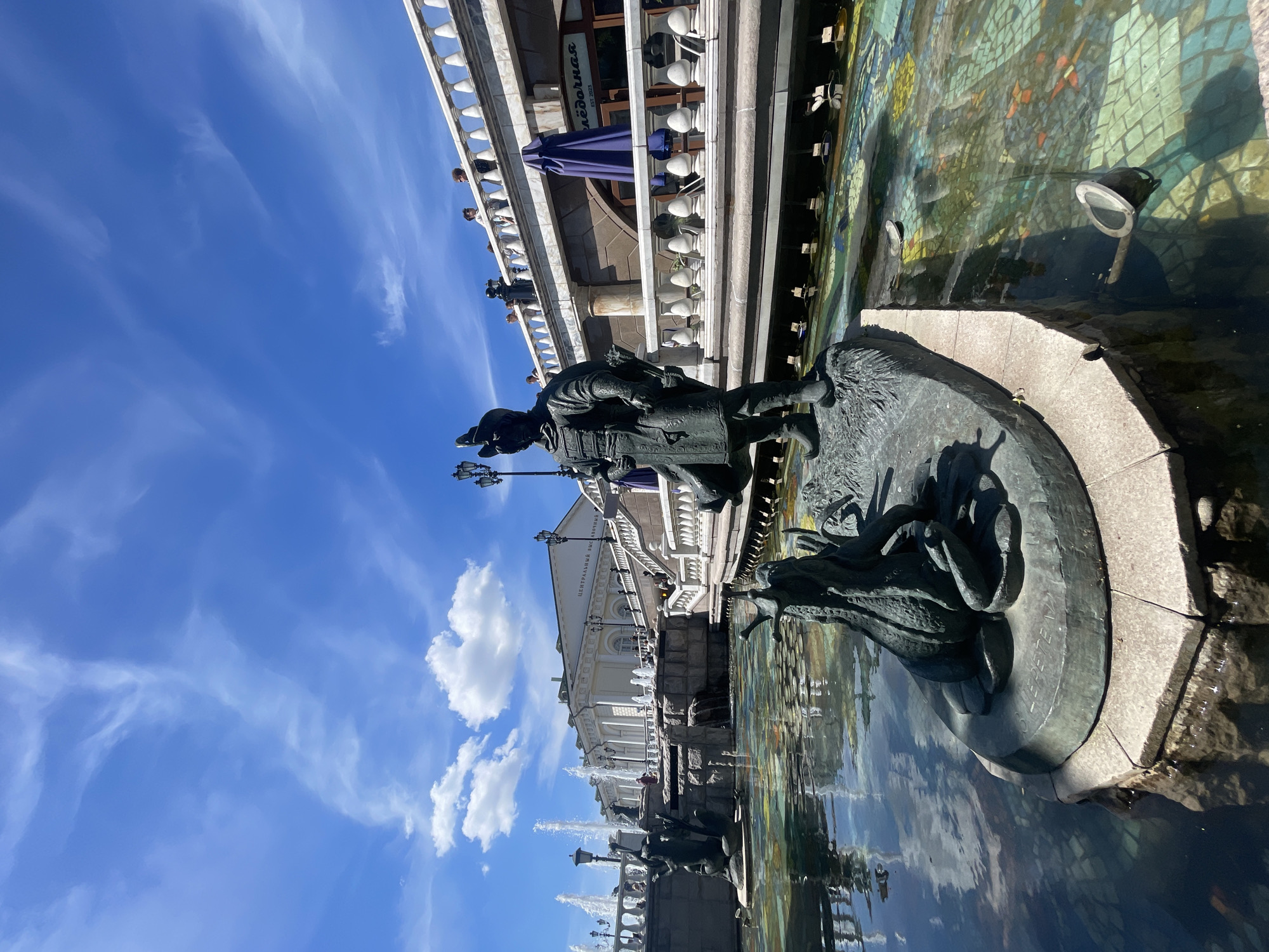 A bronze statue of a person and a creature stands in a fountain. The person is wearing a helmet and tunic, holding a spear. The creature appears to be a frog or toad. The fountain has a tiled bottom with colorful mosaic patterns of fish and flowers. In the background, a white building with columns and a grand staircase leads up to a terrace. People are visible on the terrace and on the staircase. Several fountains spray water in the distance. The sky is blue with scattered white clouds. The scene appears to be a public square or park on a sunny day.