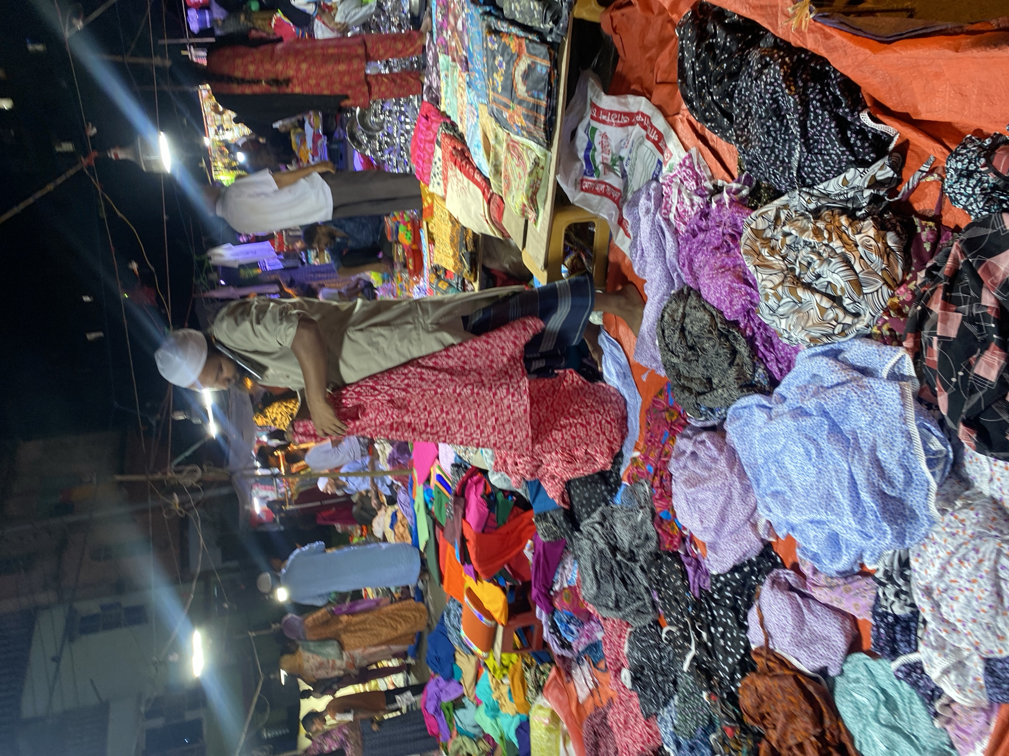 This image captures a nighttime scene at a street market in Dhaka, Bangladesh. A variety of colorful fabrics and clothing items are piled high on an orange tarpaulin, suggesting a textile market or a stall selling garments. Several people are present, appearing to be vendors and customers. One man in the foreground, wearing a white cap and a light brown shirt, is holding a red patterned fabric. Other individuals are visible in the background, some in traditional attire. The market is illuminated by artificial lights, casting a slightly hazy glow and creating light streaks across the scene. The overall impression is one of a bustling, active marketplace.